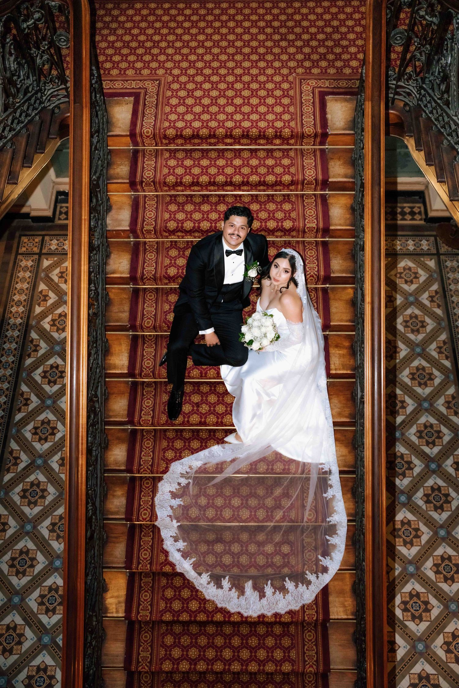 Aerial view of bride and groom sitting on grand staircase with red carpet and ornate tile floor.