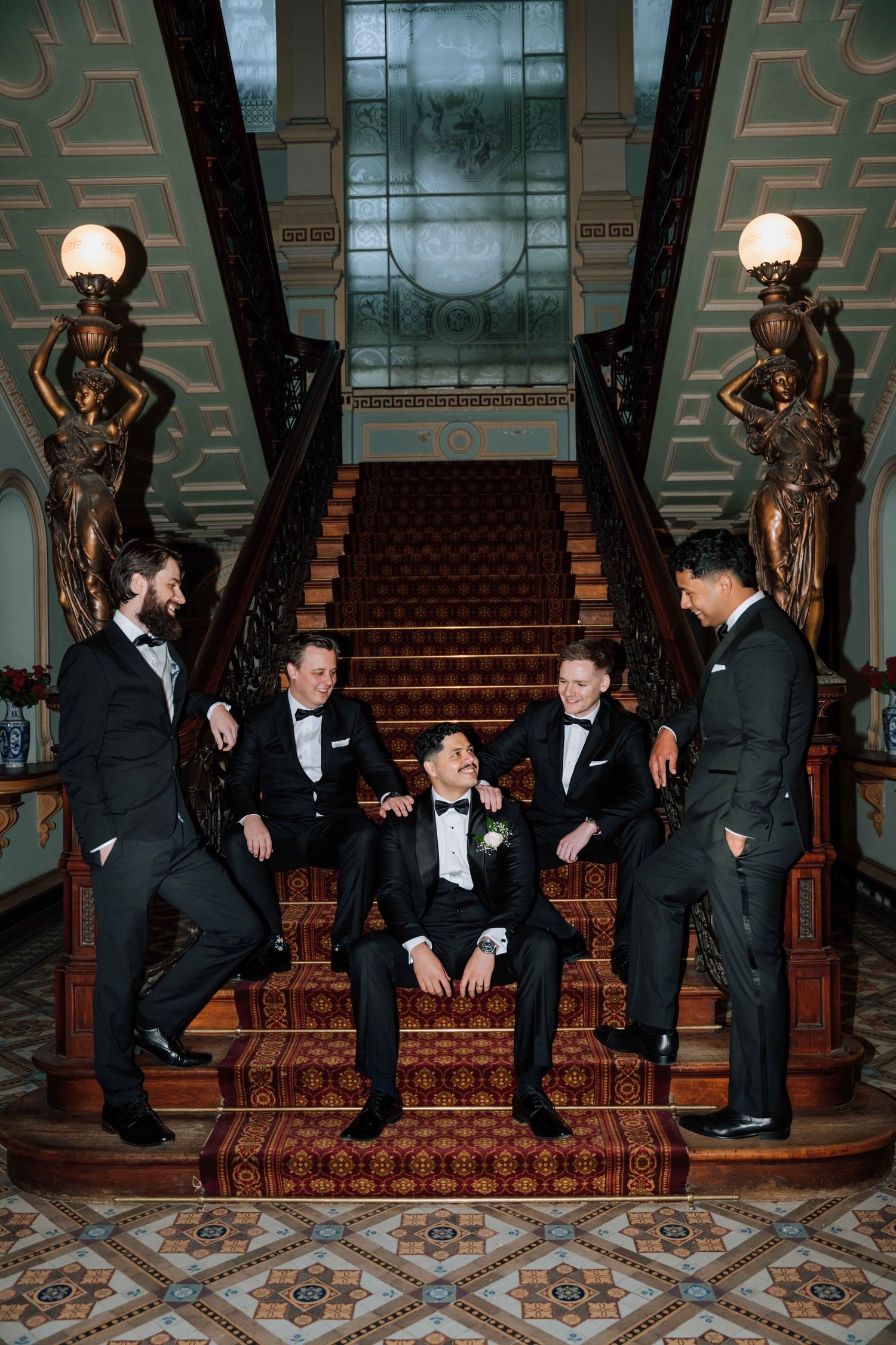 Groom and groomsmen in black tuxedos laughing together on ornate staircase in elegant wedding venue.