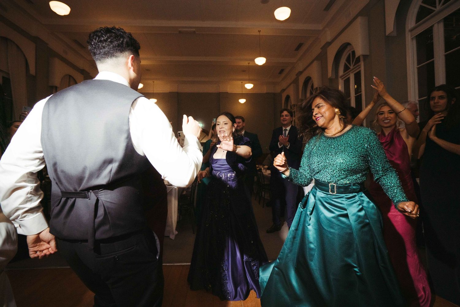 Guests dance joyfully at wedding reception including woman in teal gown and another in navy dress.