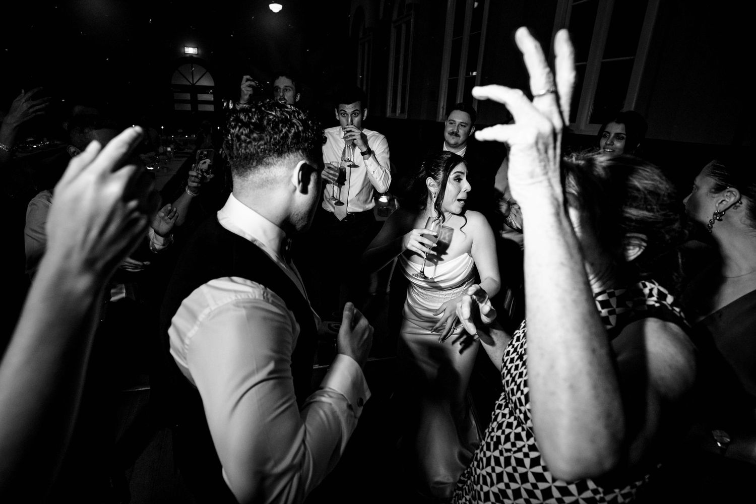 Black and white photo of bride and groom dancing with cheering guests at lively wedding reception.
