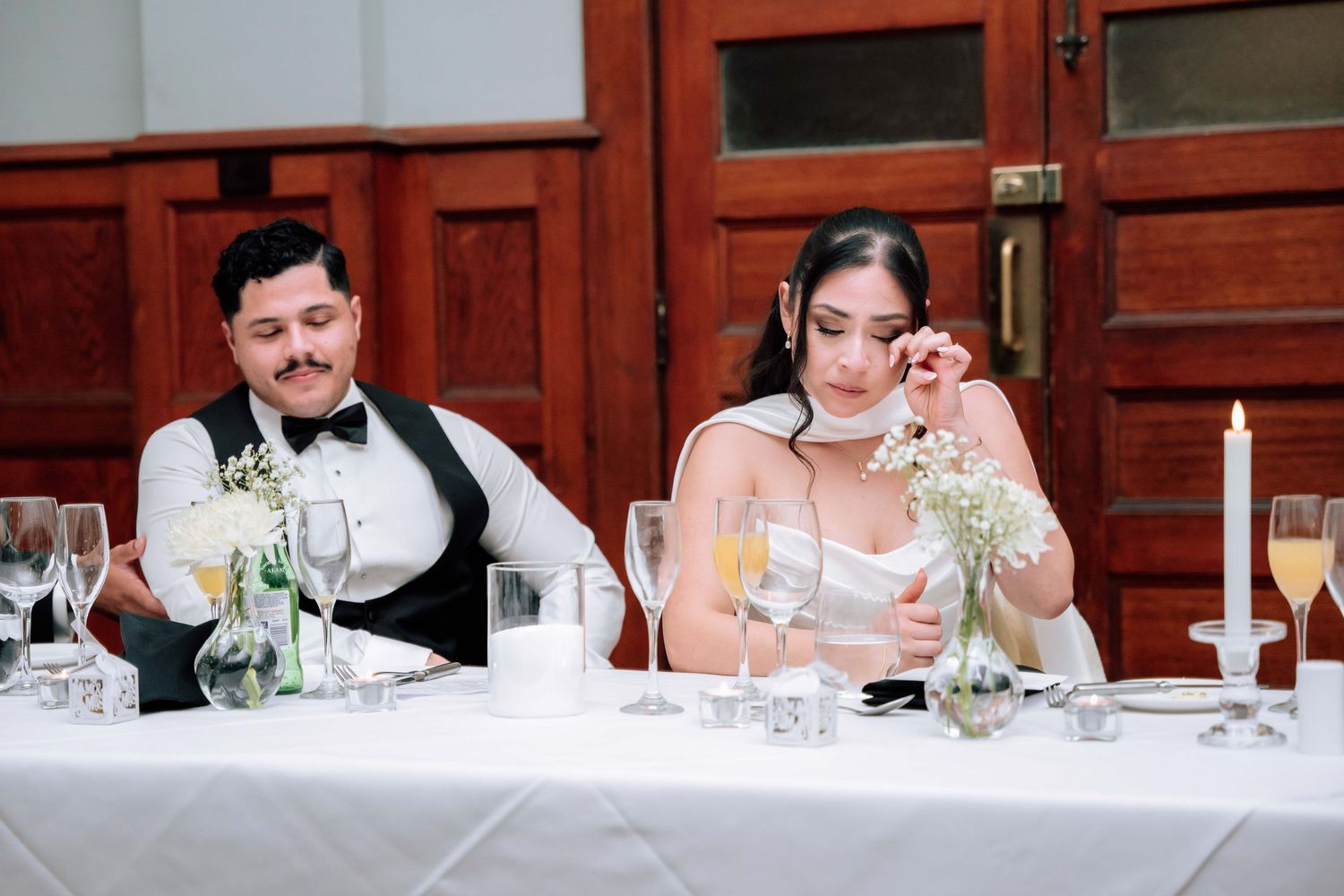 Bride wiping tears while groom smiles at sweetheart table during wedding reception speeches.