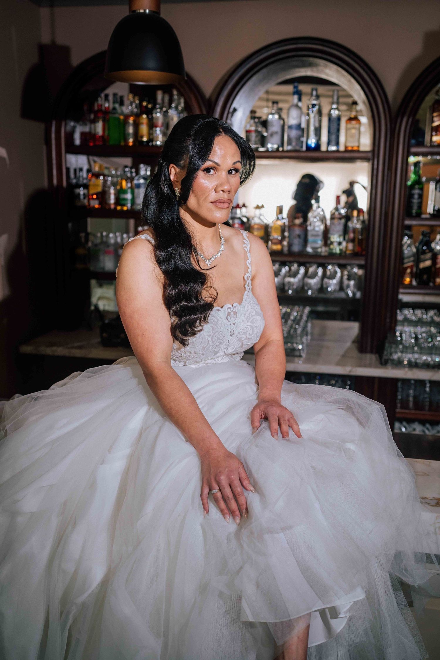 A bride in a layered white tulle wedding gown sits gracefully at a bar with bottle-lined shelves.