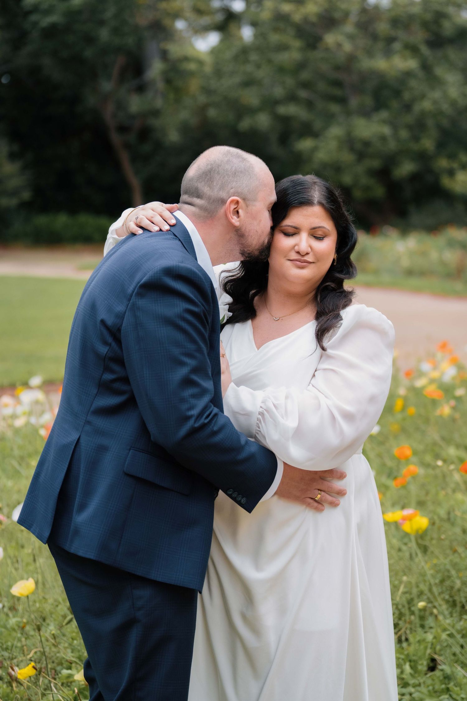 Groom tenderly kisses bride on the cheek while embracing her in a colorful garden on their wedding day.