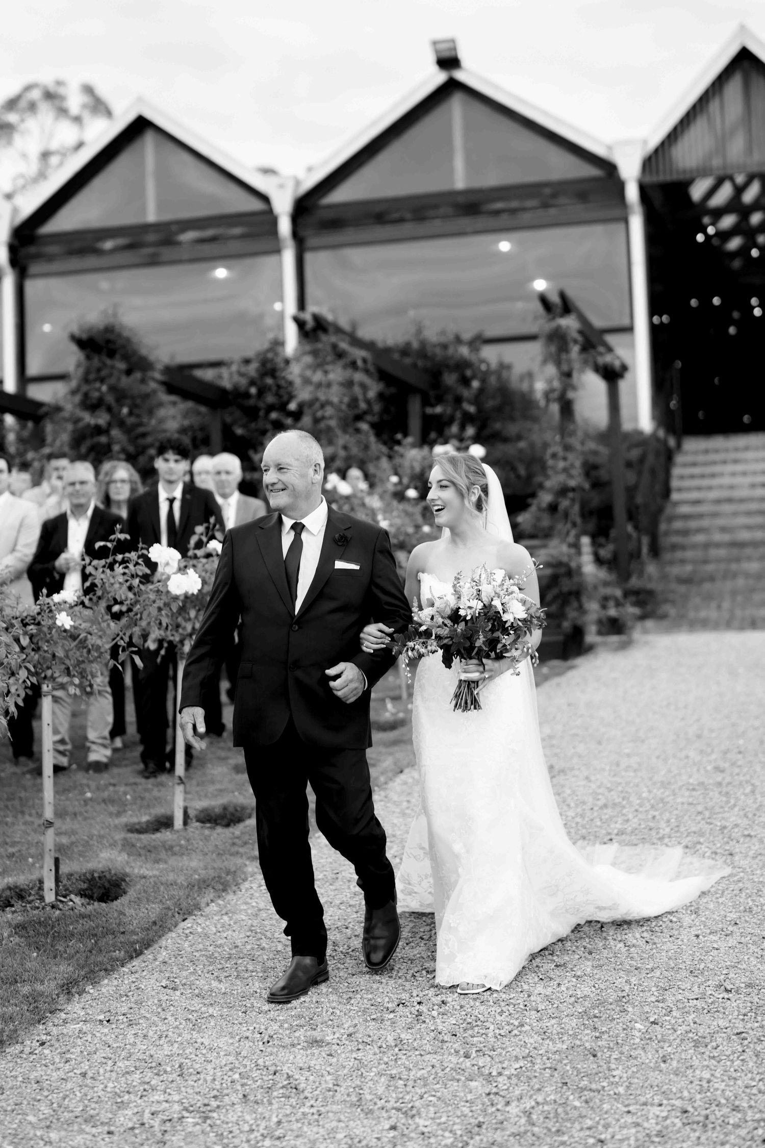 Black and white photo of a smiling bride walking down the aisle with her father at an outdoor wedding.