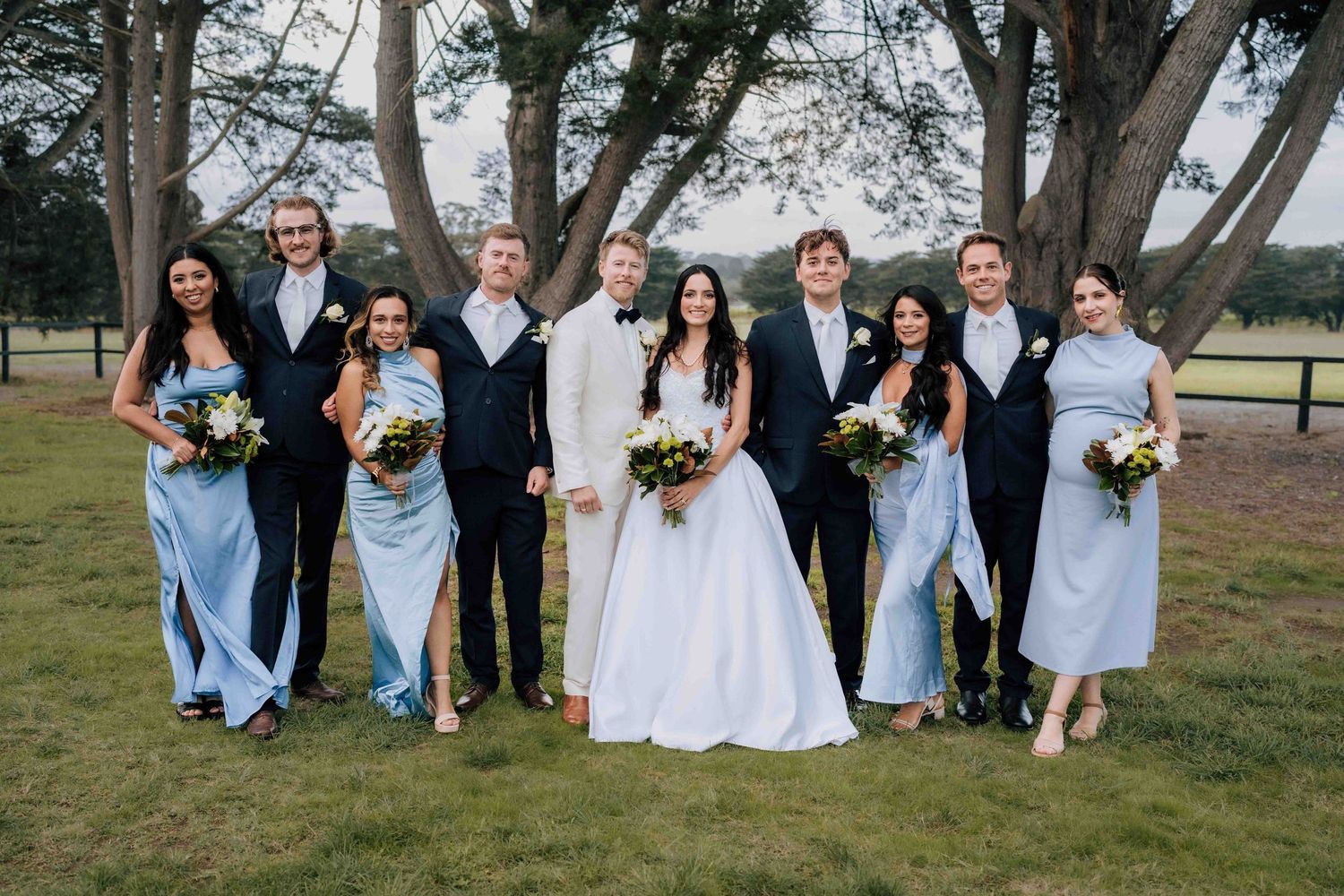 Wedding party posing outdoors in blue bridesmaid dresses and dark suits with bride and groom in white.
