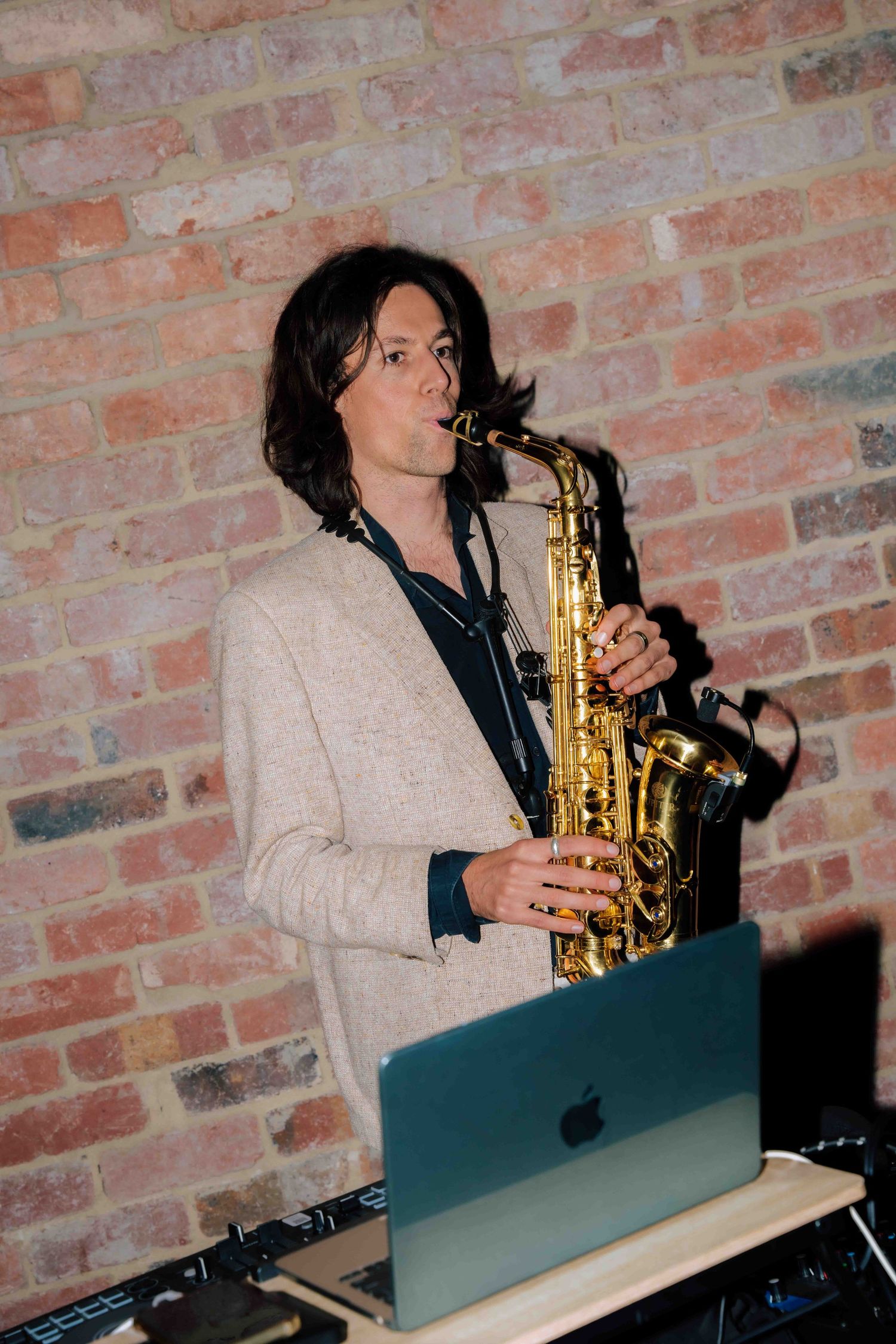 Musician playing a gold saxophone at a wedding reception, wearing a beige blazer with a MacBook on the DJ table.