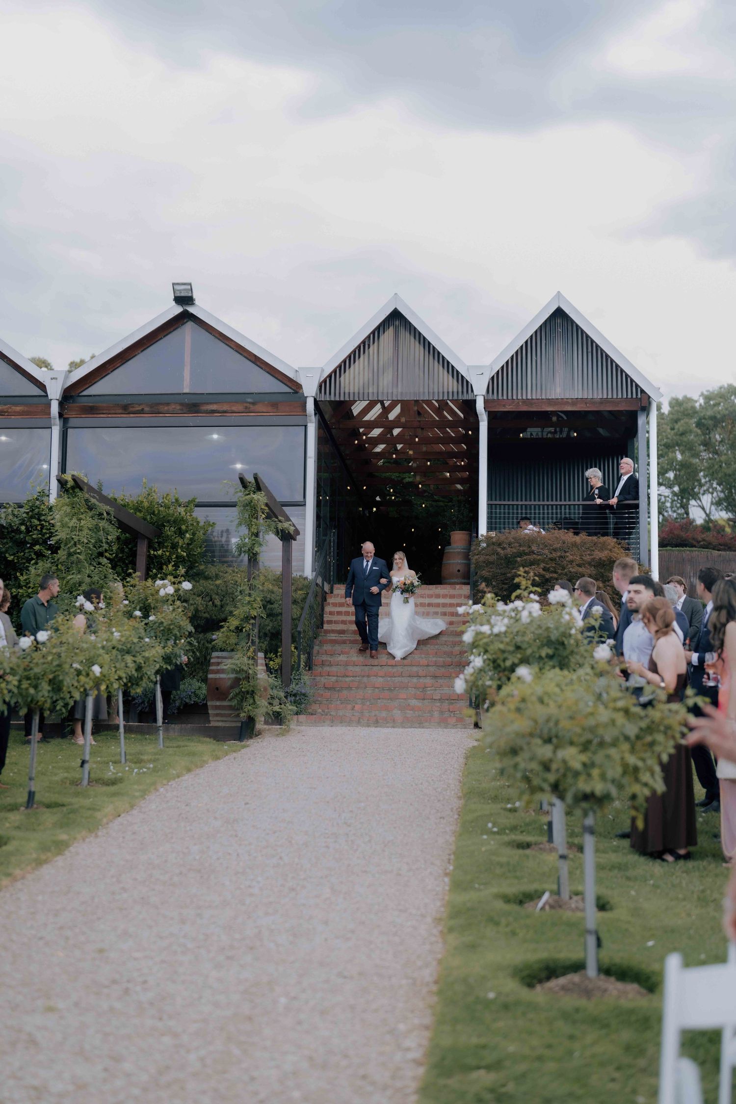 Bride and father descend steps from barn venue toward outdoor wedding ceremony aisle with guests.