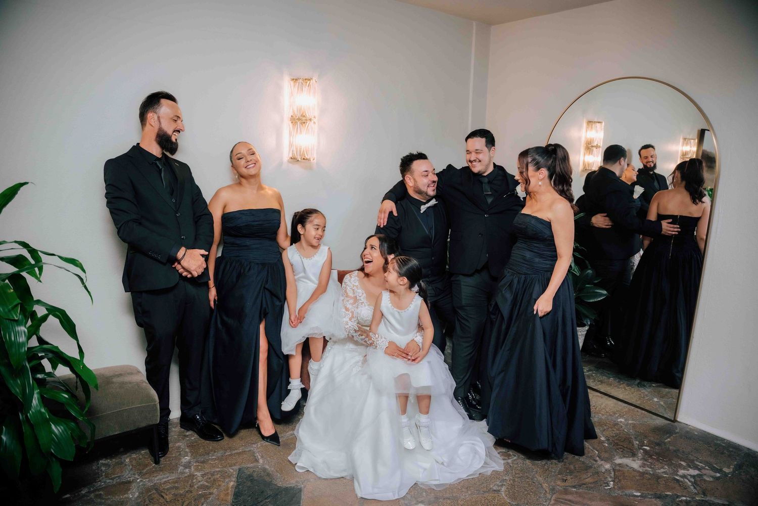 Joyful wedding party laughing together in black formal wear with bride and flower girls in white.