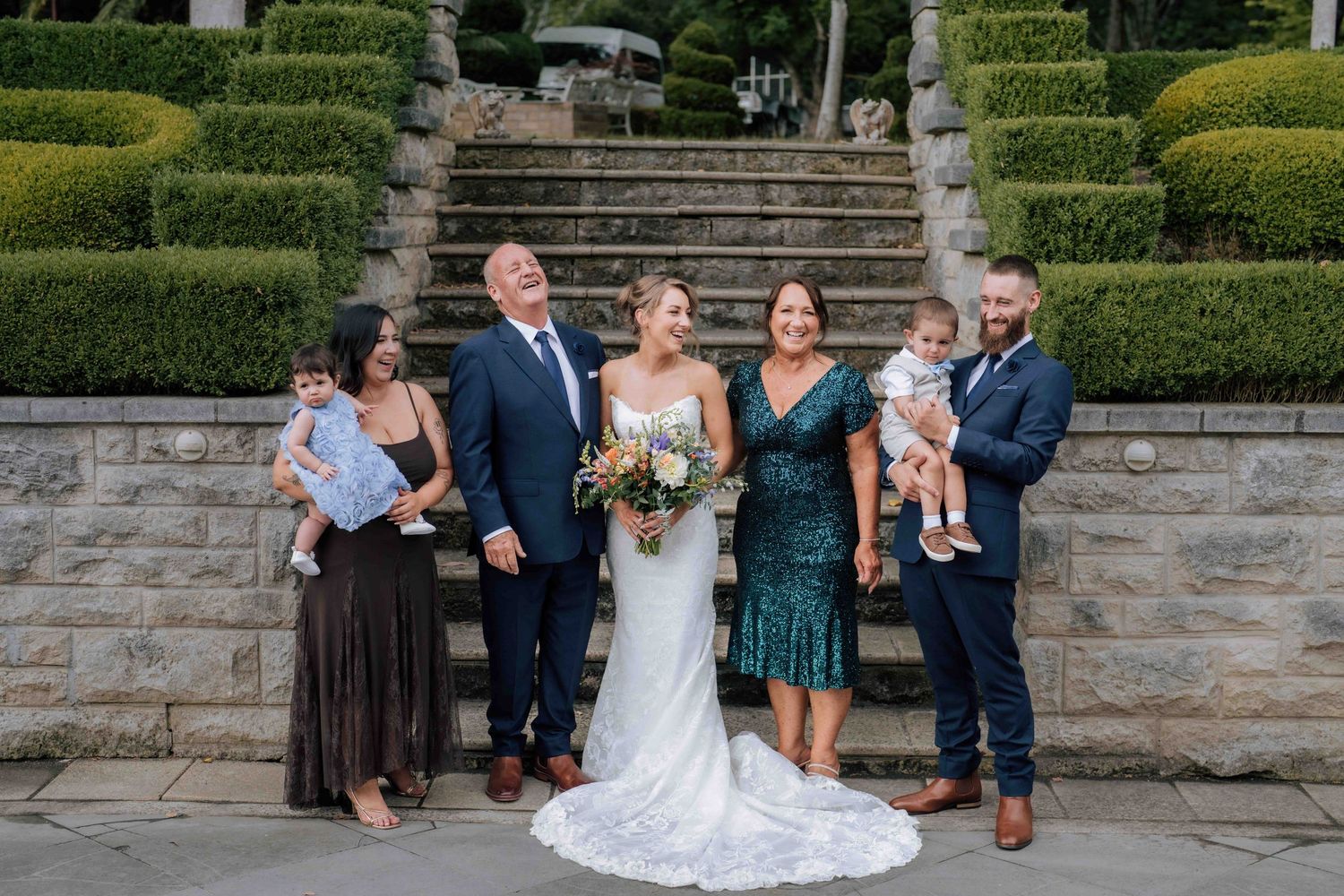 Wedding party group photo with bride and groom flanked by family members holding babies on stone garden steps.