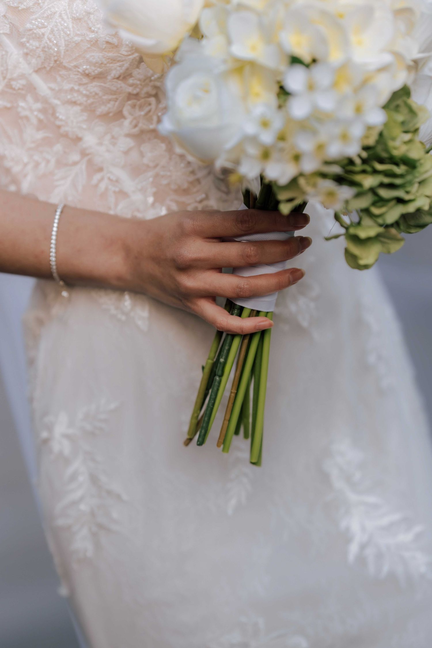 Bride holding white floral bouquet with green stems in lace wedding dress with pearl bracelet.