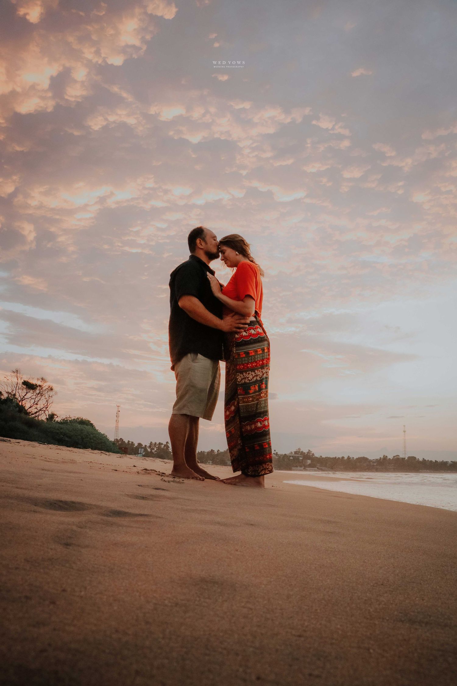 Couple sharing romantic moment on beach at sunset under dramatic pink cloudy sky.