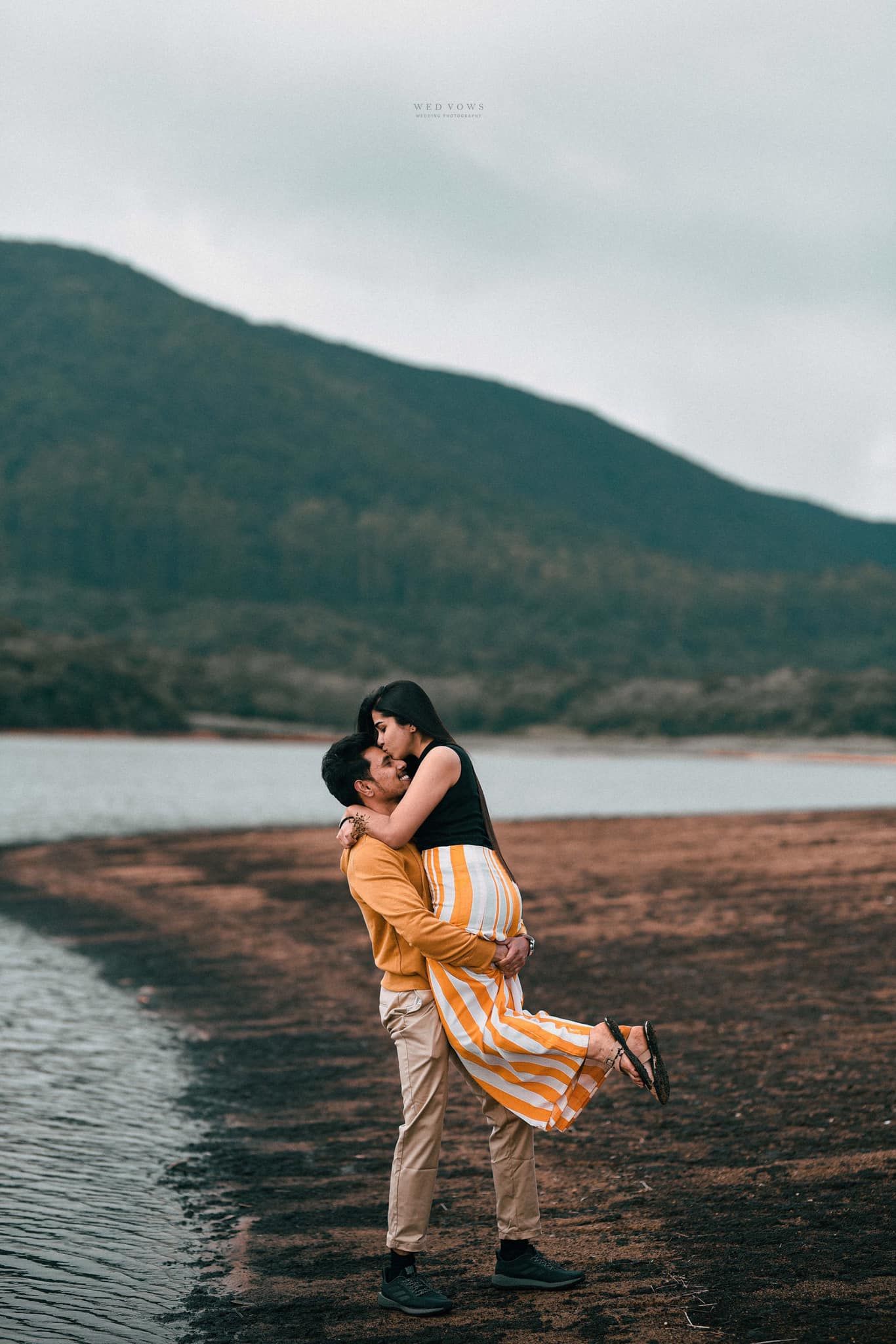 Man lifting woman in orange striped dress near a mountain lake in a romantic outdoor photo shoot.