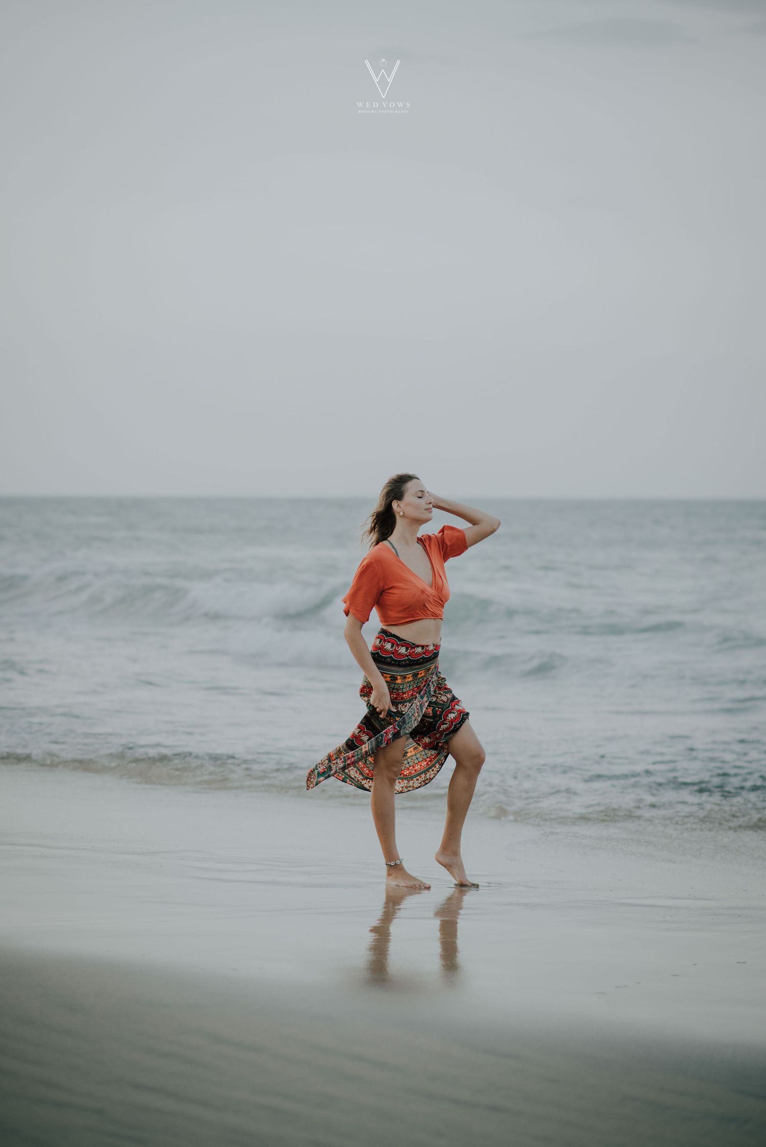 Woman in orange crop top and floral skirt dancing barefoot on a beach shoreline.