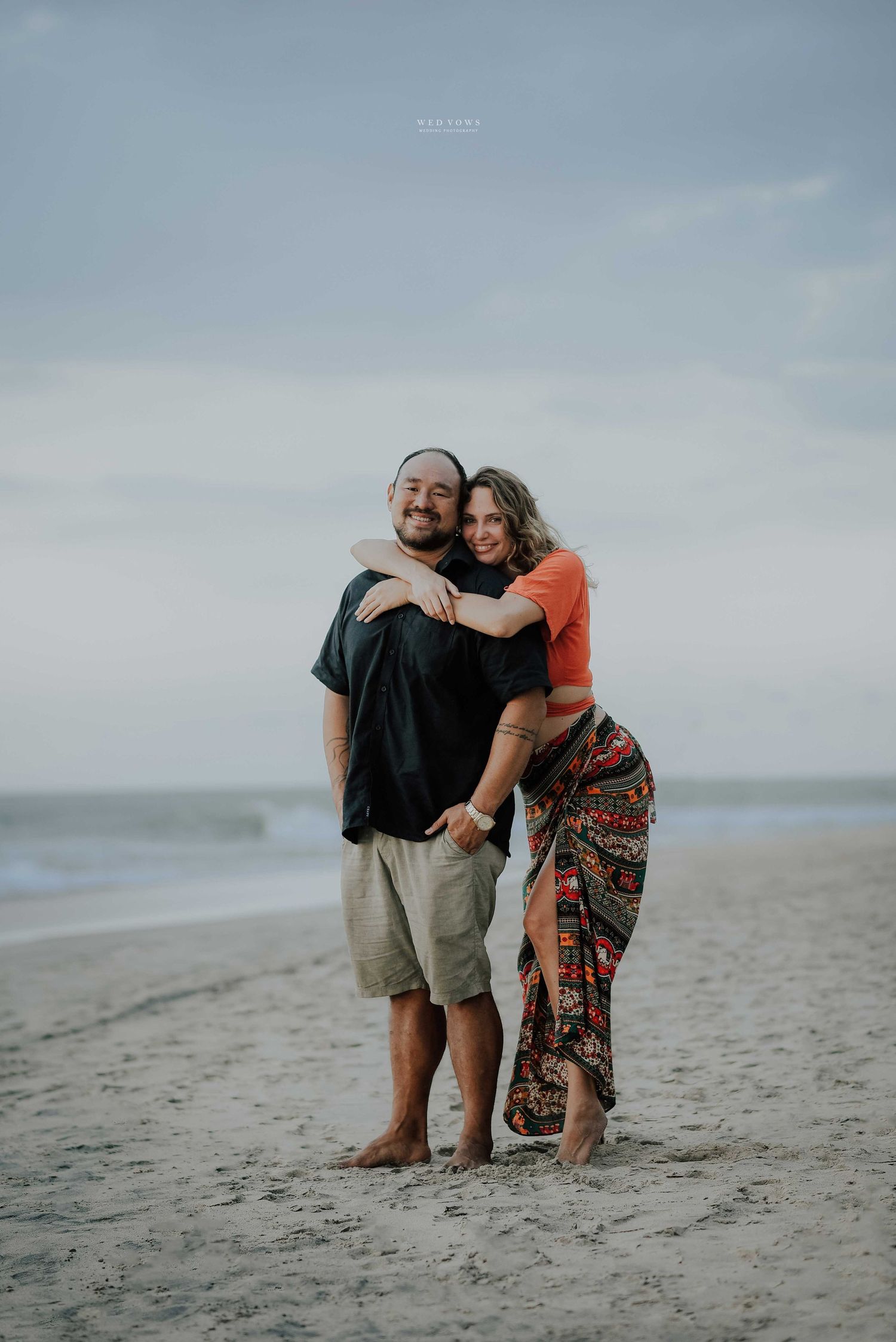 Smiling couple hugging on sandy beach, woman in orange top and patterned skirt, ocean behind.