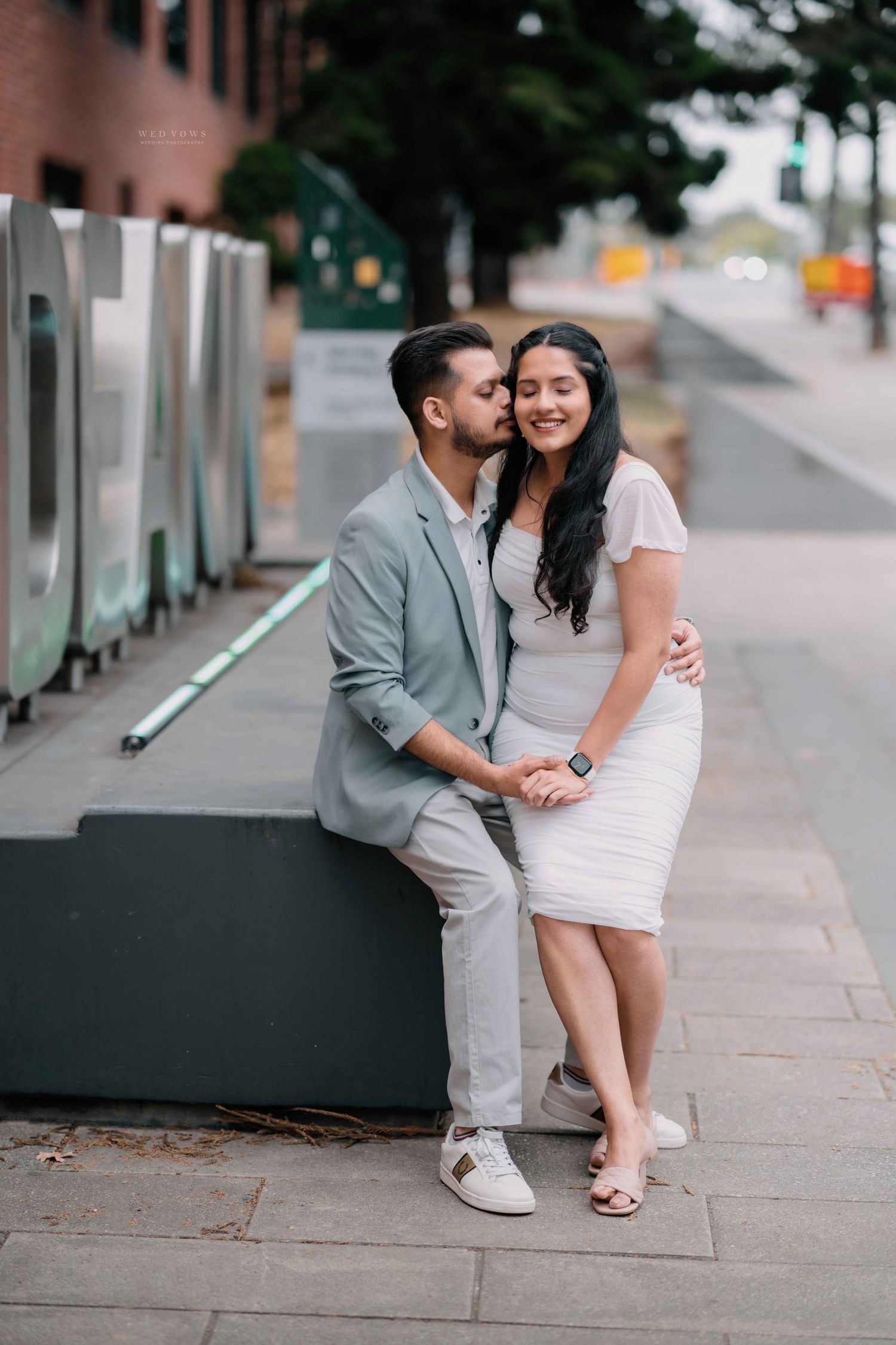 Man in grey blazer kisses woman in white dress on the cheek while seated on an urban bench.