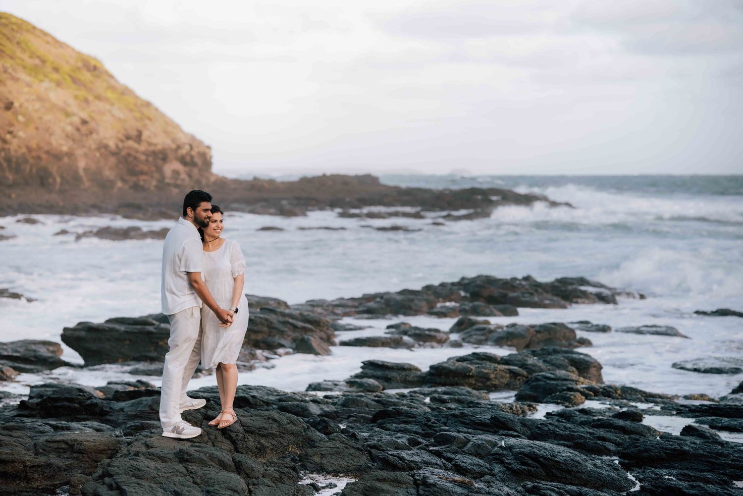 Couple in white outfits embracing on rocky shoreline with ocean waves behind them.