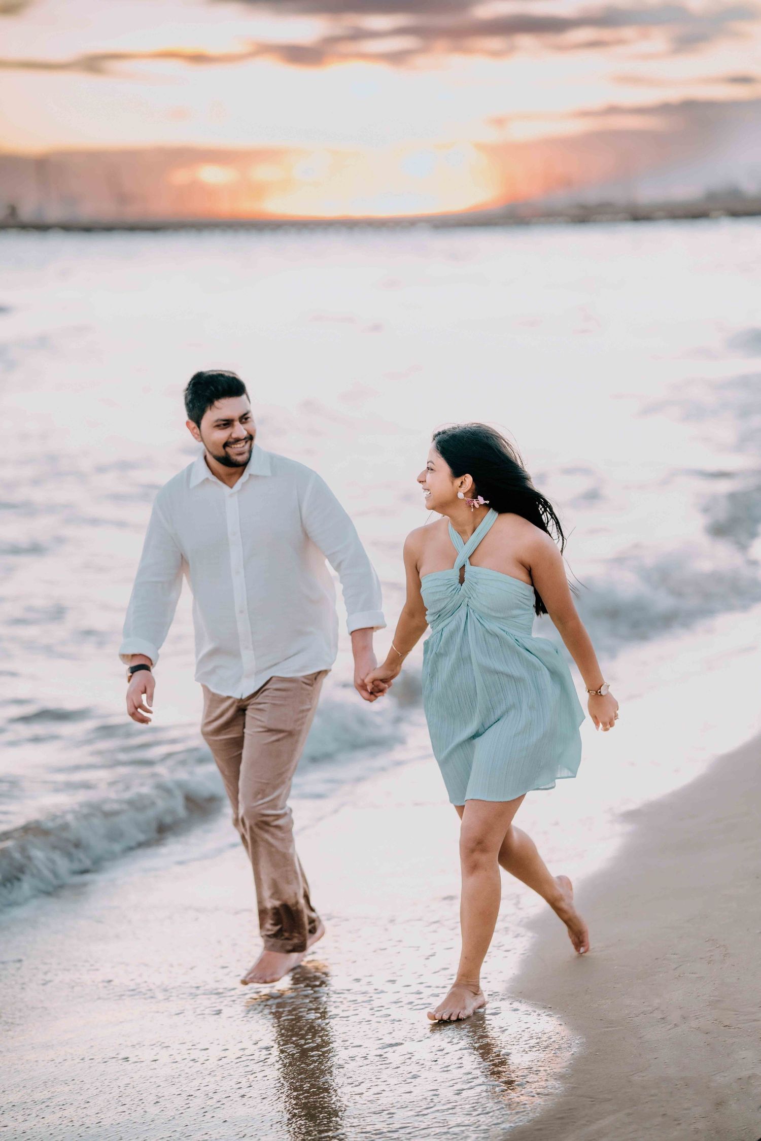 Joyful couple holding hands walking along beach at vibrant sunset, woman in light blue halter dress.