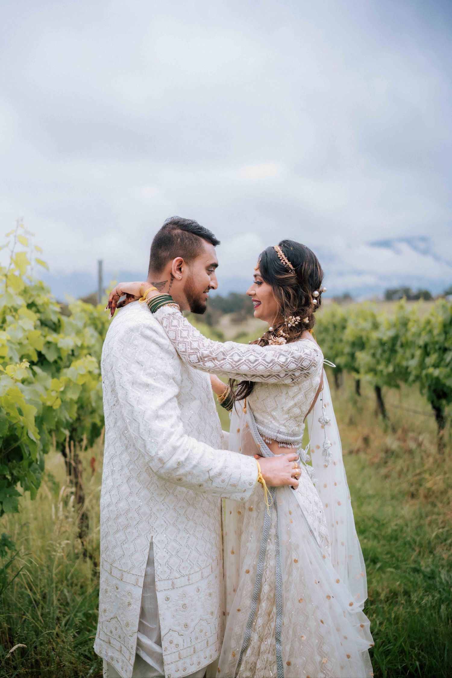 Indian couple in matching white embroidered wedding outfits embracing in a vineyard setting.