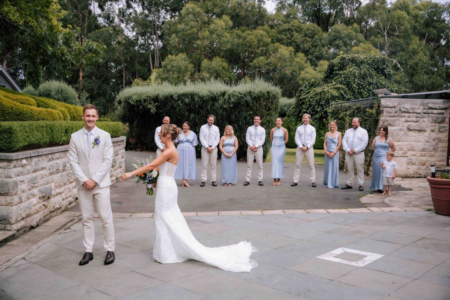 Groom smiles as bride approaches in white gown during first look on stone patio with bridal party.