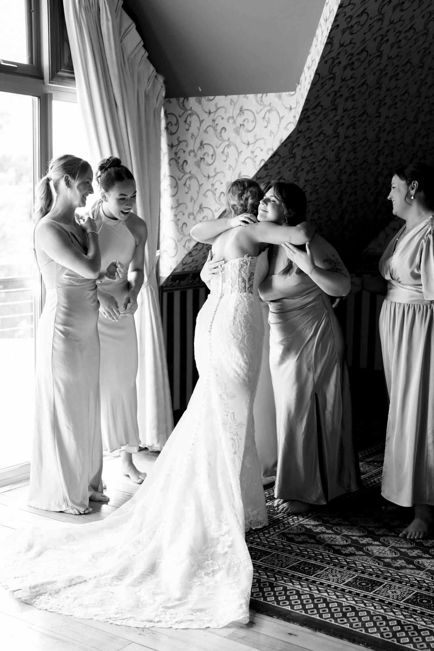 Black and white photo of a bride in a lace gown sharing heartfelt hugs with her bridesmaids before the ceremony.