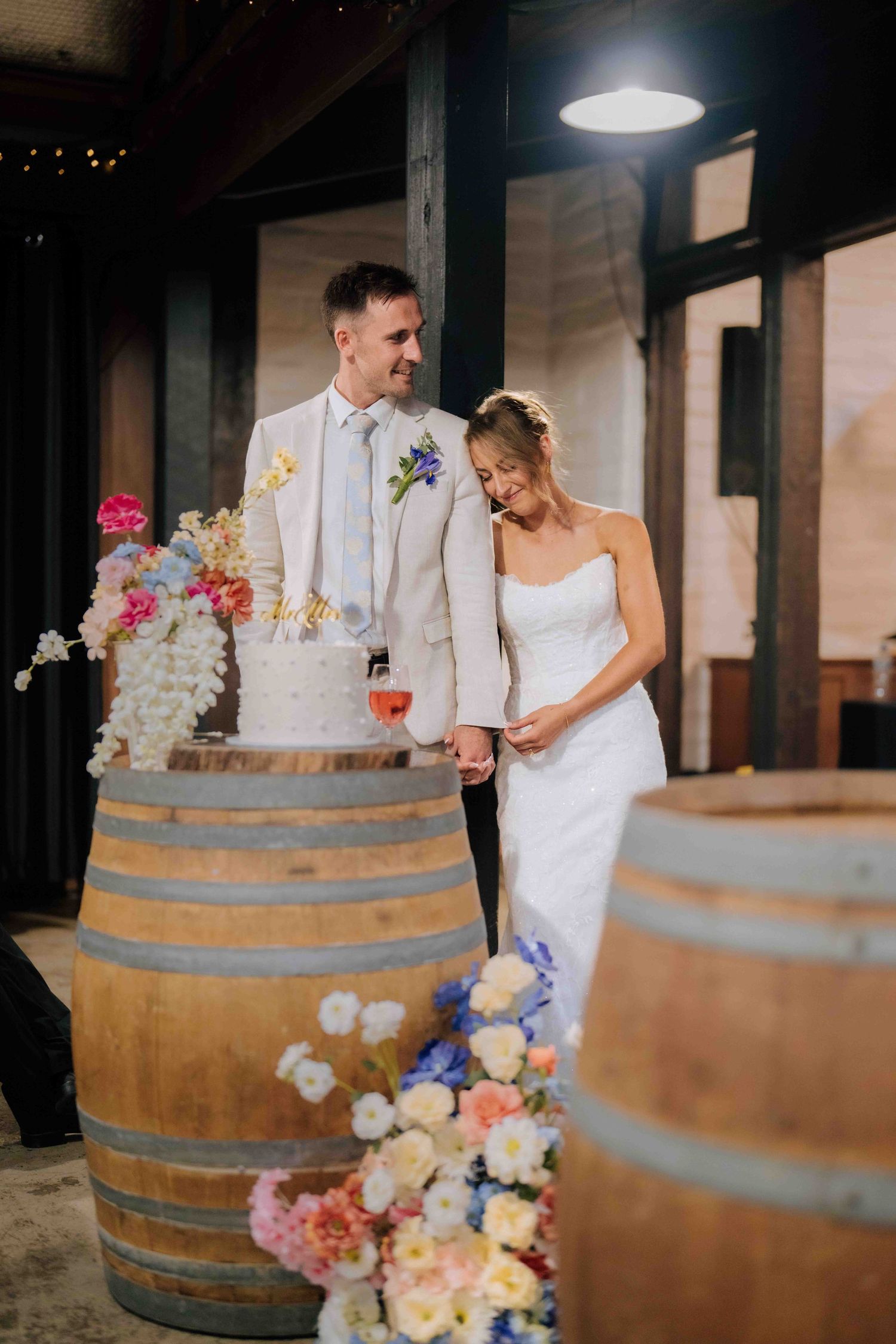 Bride and groom stand beside floral wedding cake displayed on rustic wooden wine barrels.