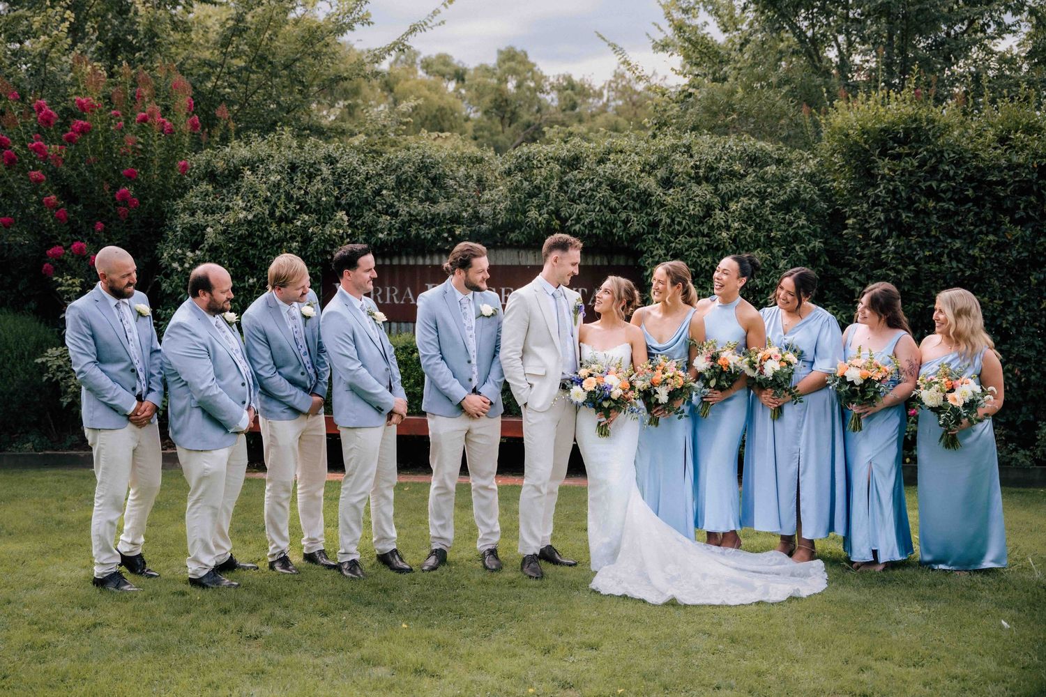 Wedding party in coordinated blue and cream outfits poses on garden lawn with bouquets.