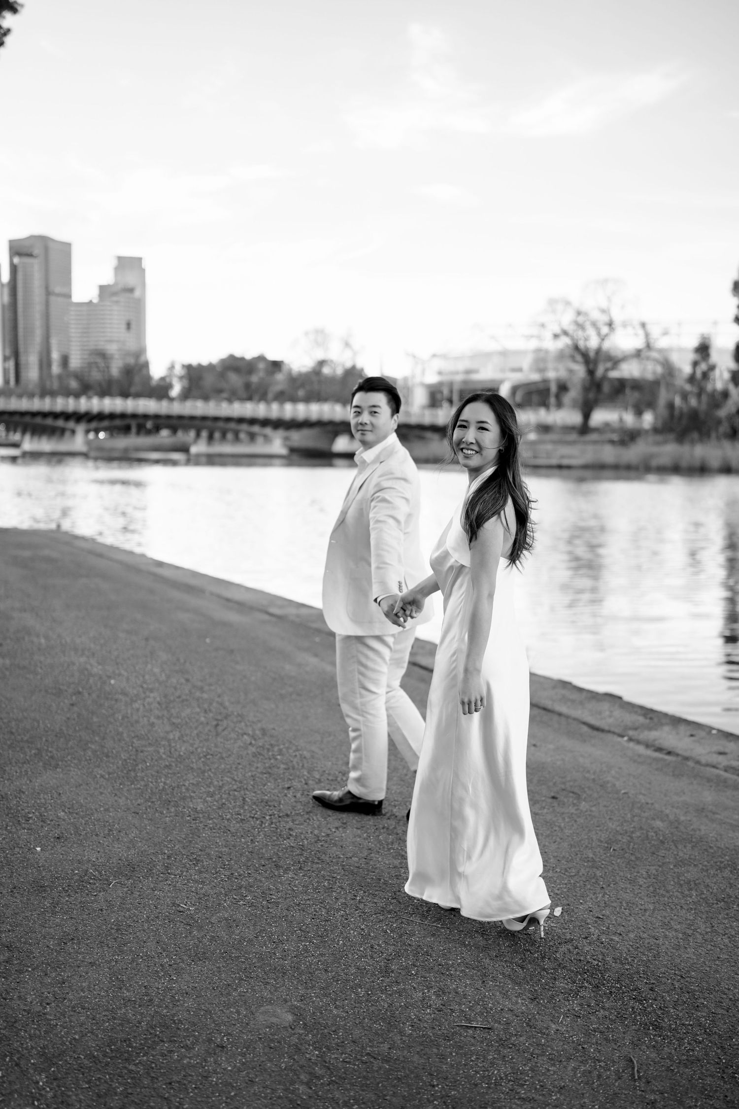 Black and white image of Asian couple in white wedding attire holding hands while walking by a city river.