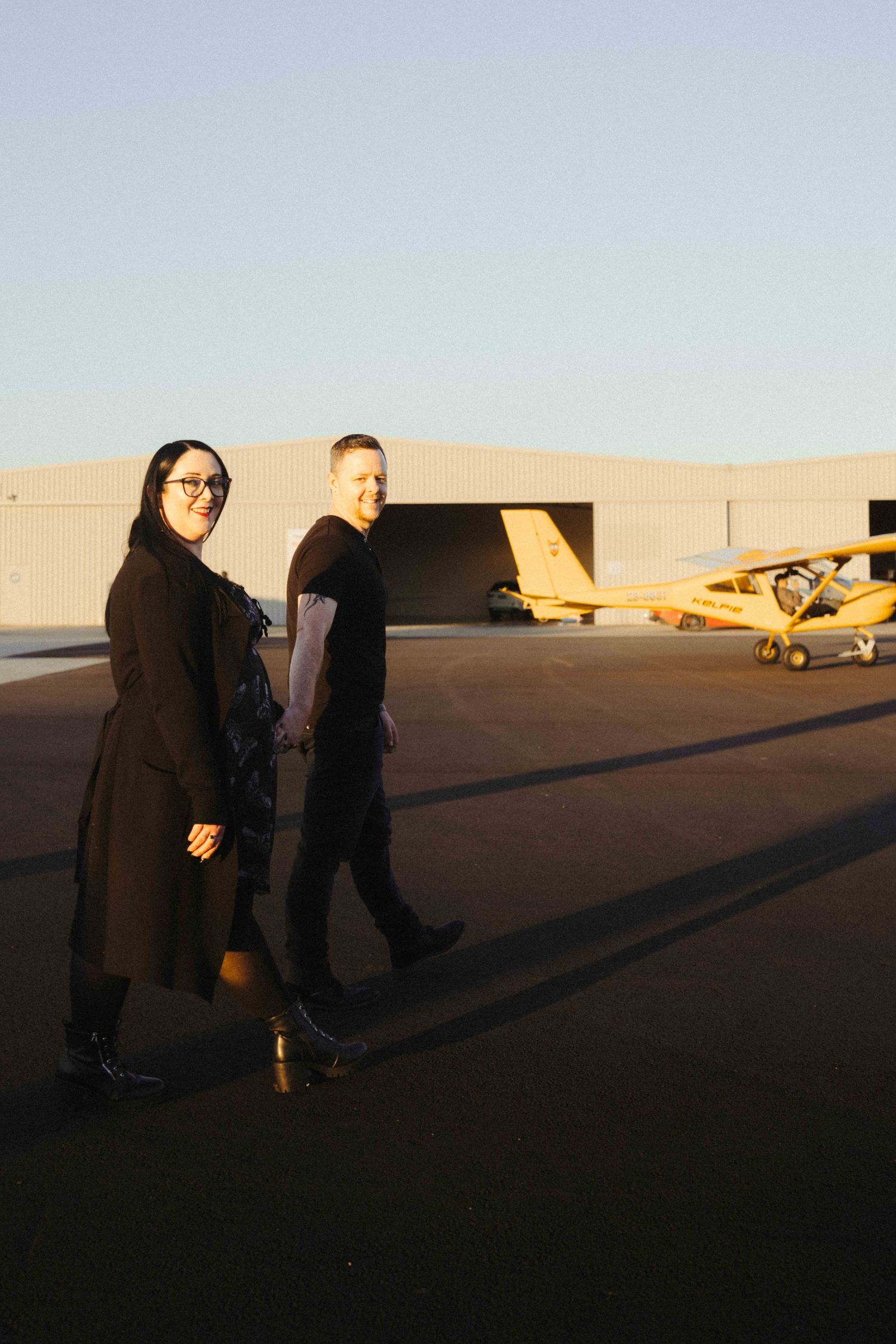 Couple walking hand in hand on an airfield tarmac with a yellow plane and hangar in the background.