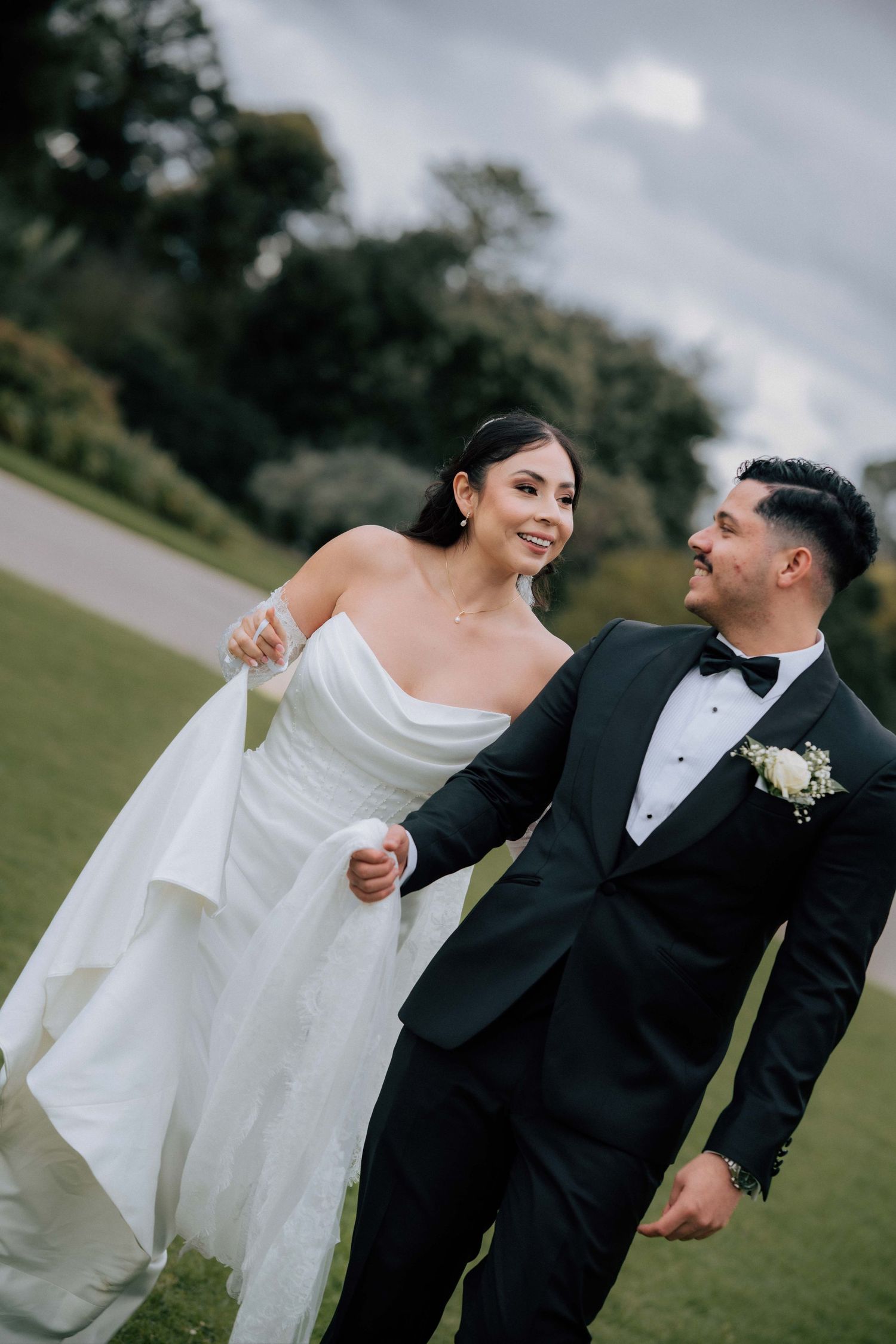 Bride and groom walk hand in hand across green lawn, smiling at each other in elegant wedding attire.