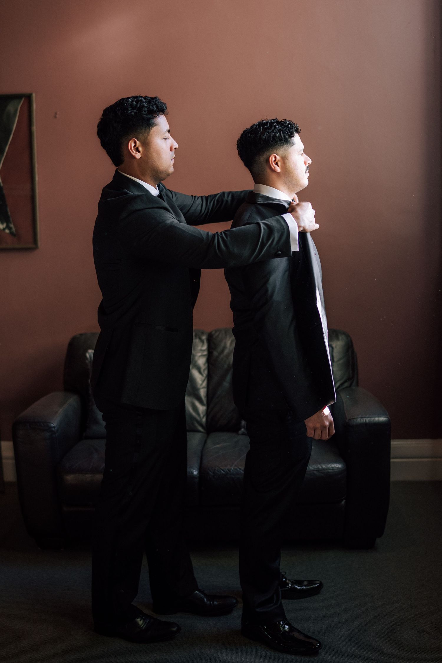 Two men in black suits, one helping the other adjust his white tie, standing in a warmly lit room with a leather sofa.