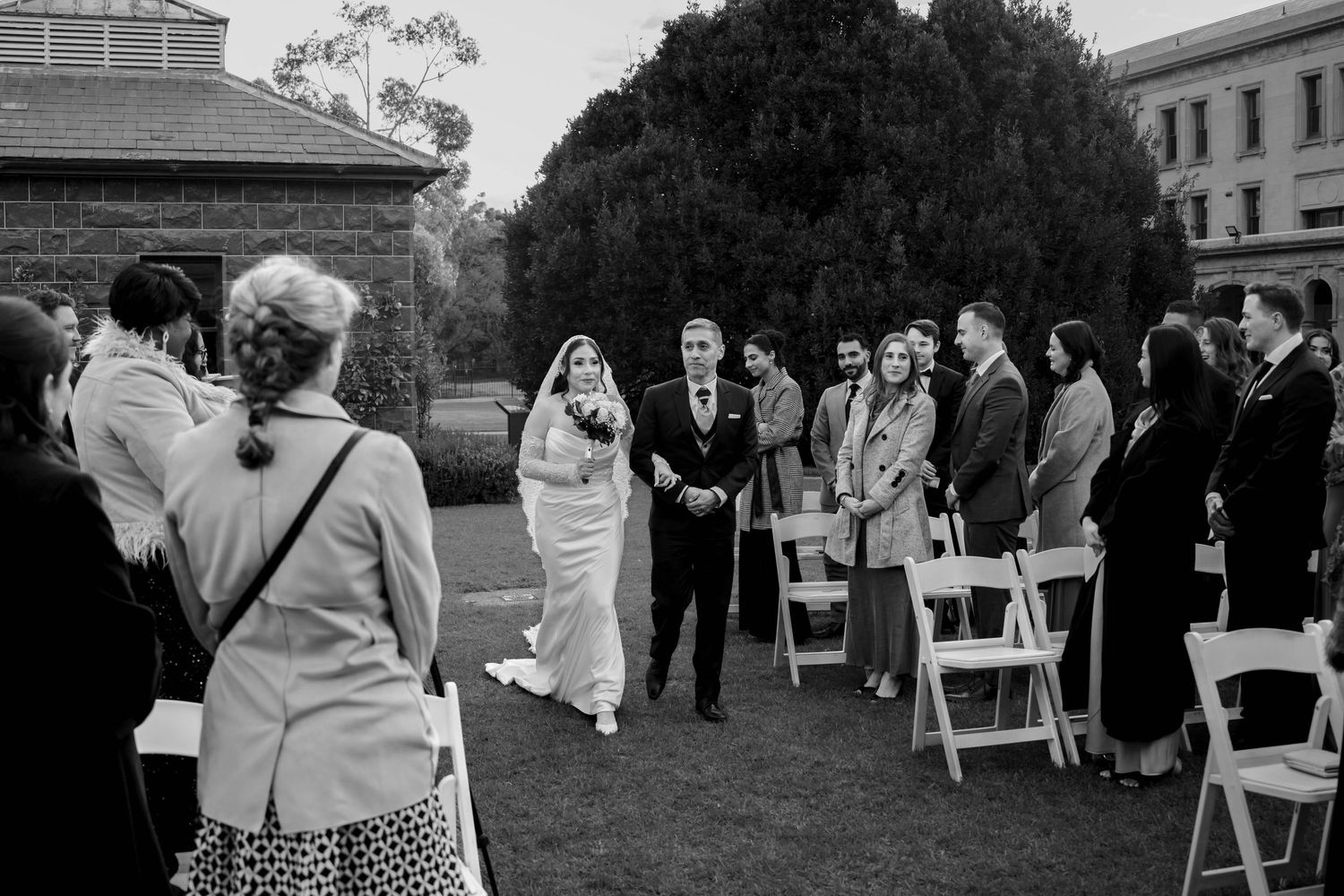 Bride walking down outdoor aisle with escort past seated guests at garden wedding ceremony.