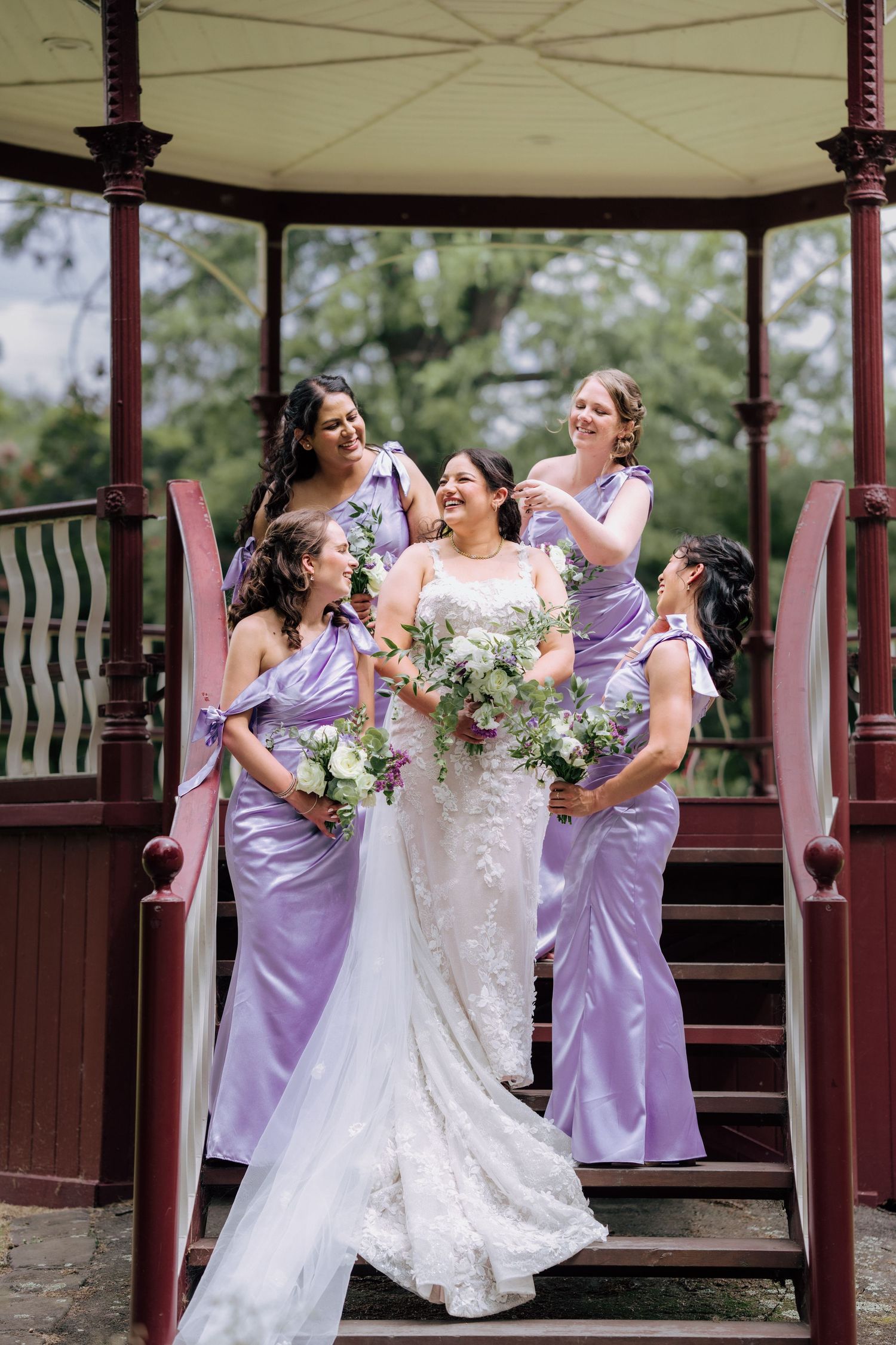 Bride laughing with bridesmaids in lavender gowns on red gazebo staircase.