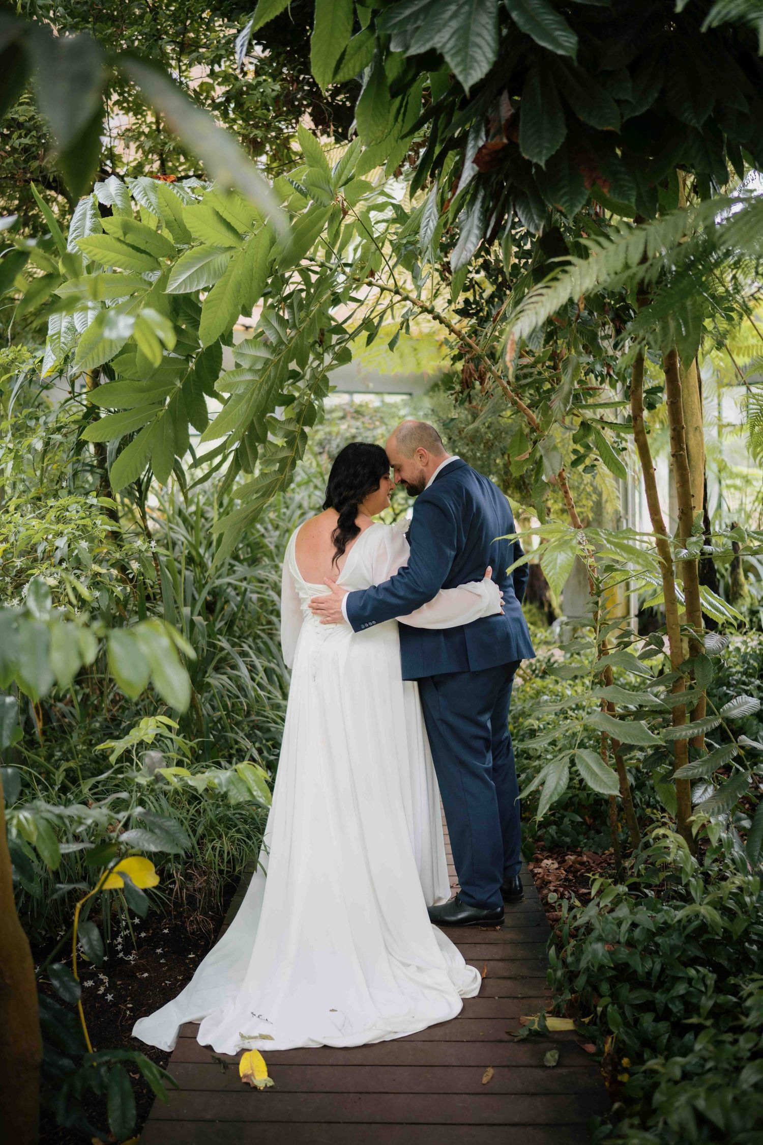 Bride and groom sharing a romantic embrace on a wooden walkway surrounded by lush tropical greenhouse foliage.