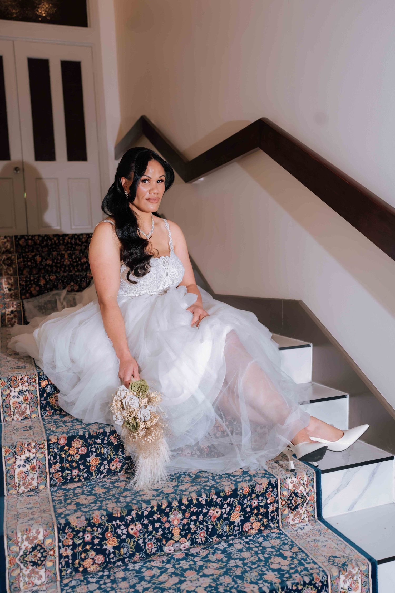 Bride in white tulle gown sitting on ornate staircase holding feather bouquet.