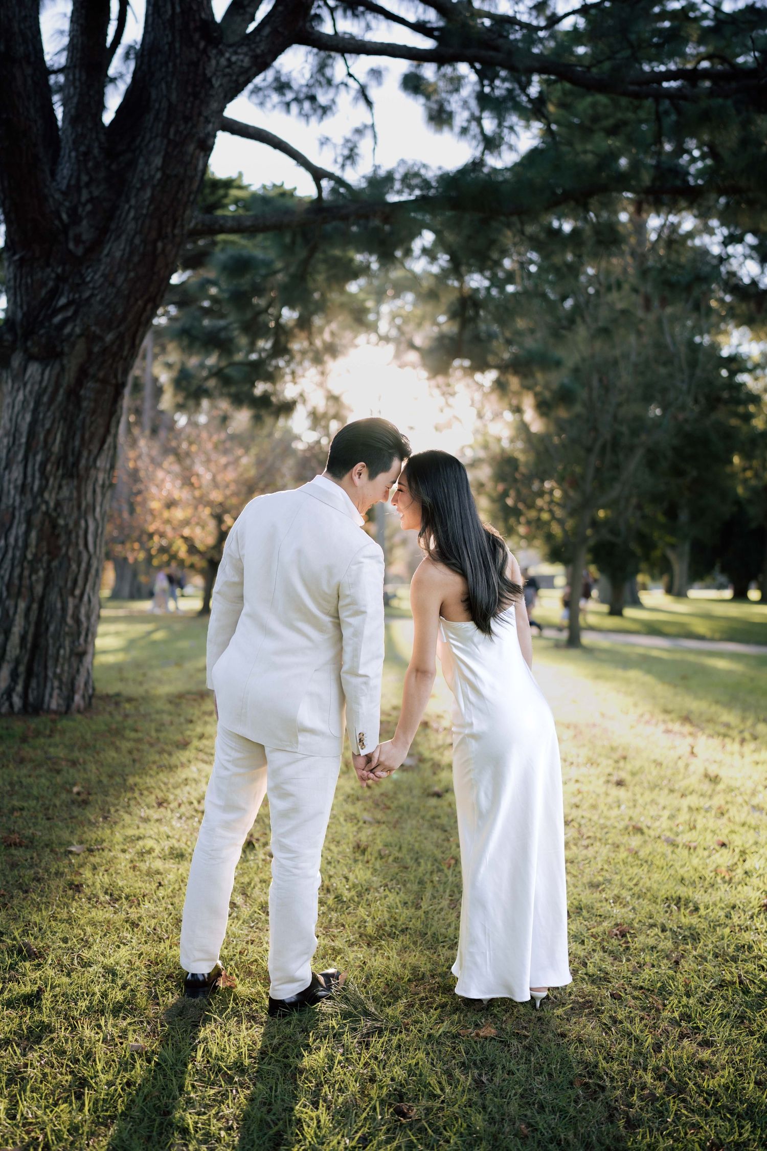 Couple in white attire holding hands and sharing a tender moment on a sunlit grassy park lawn.