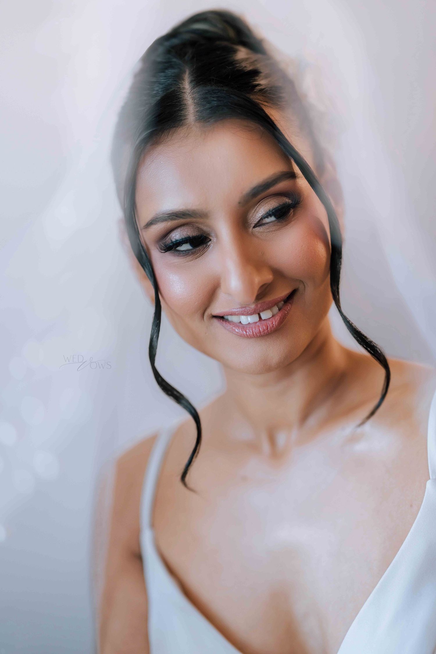 Radiant bride smiling softly under a sheer veil with elegant updo and smoky eye makeup.