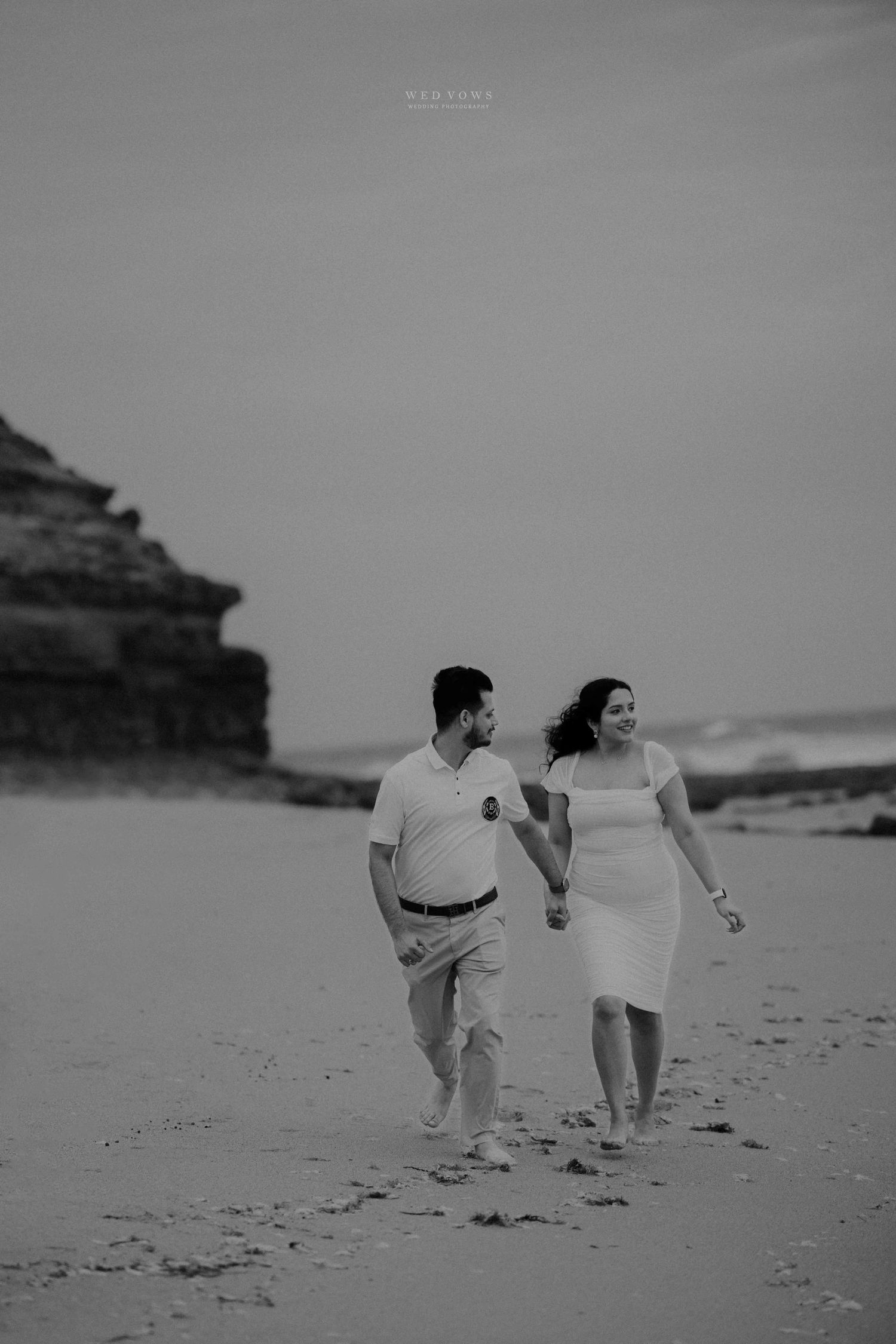 Black and white photo of couple walking hand in hand along a scenic beach shoreline.
