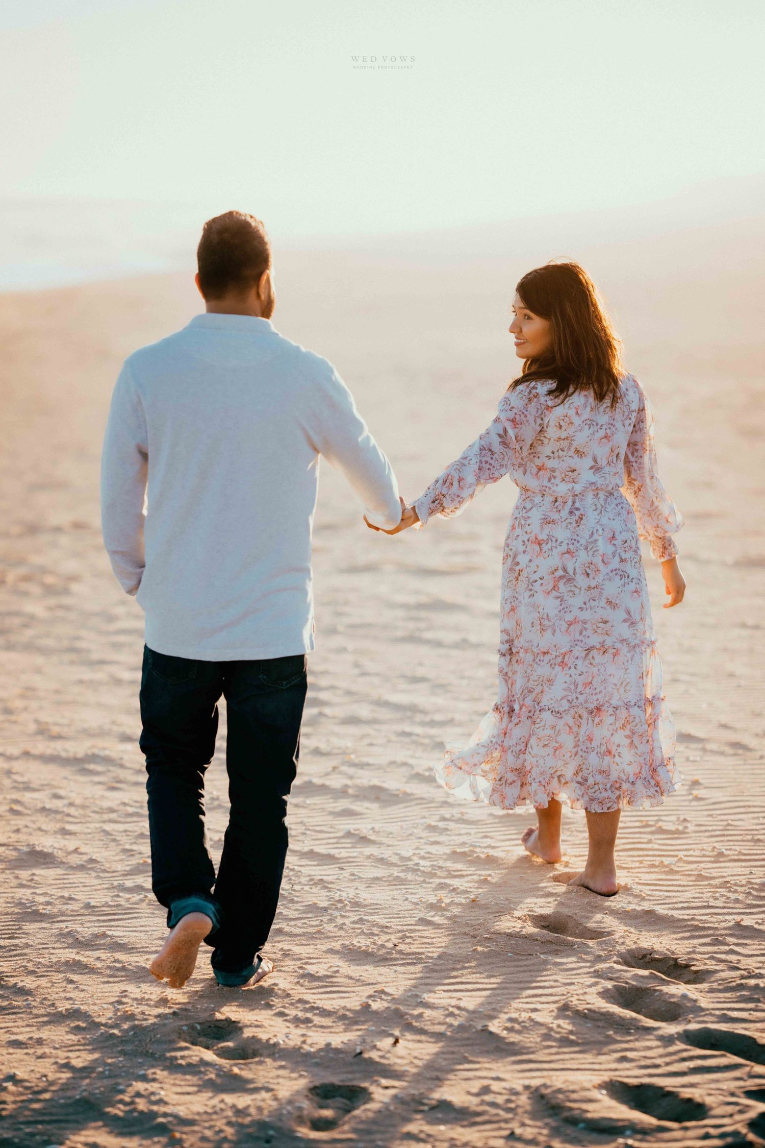 Couple walking barefoot on sandy beach at golden hour, woman in floral dress holding mans hand.