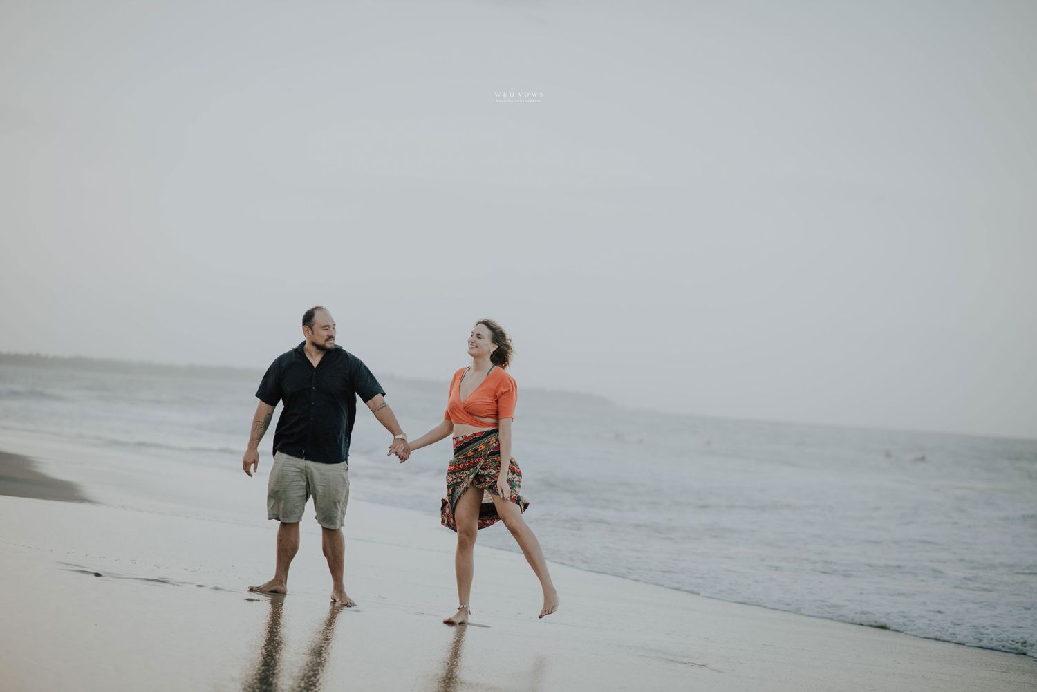 A happy couple holds hands while walking barefoot along a scenic beach with calm ocean waves nearby.