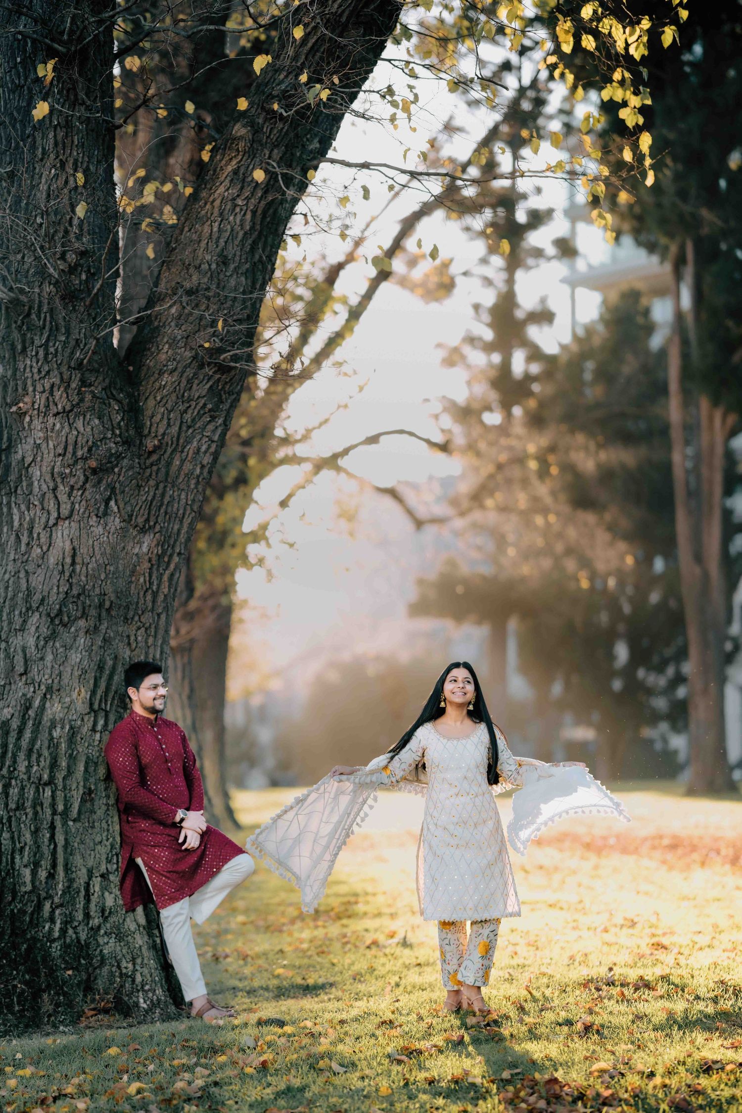 Woman in white salwar twirling near a large tree as man watches in autumn park.