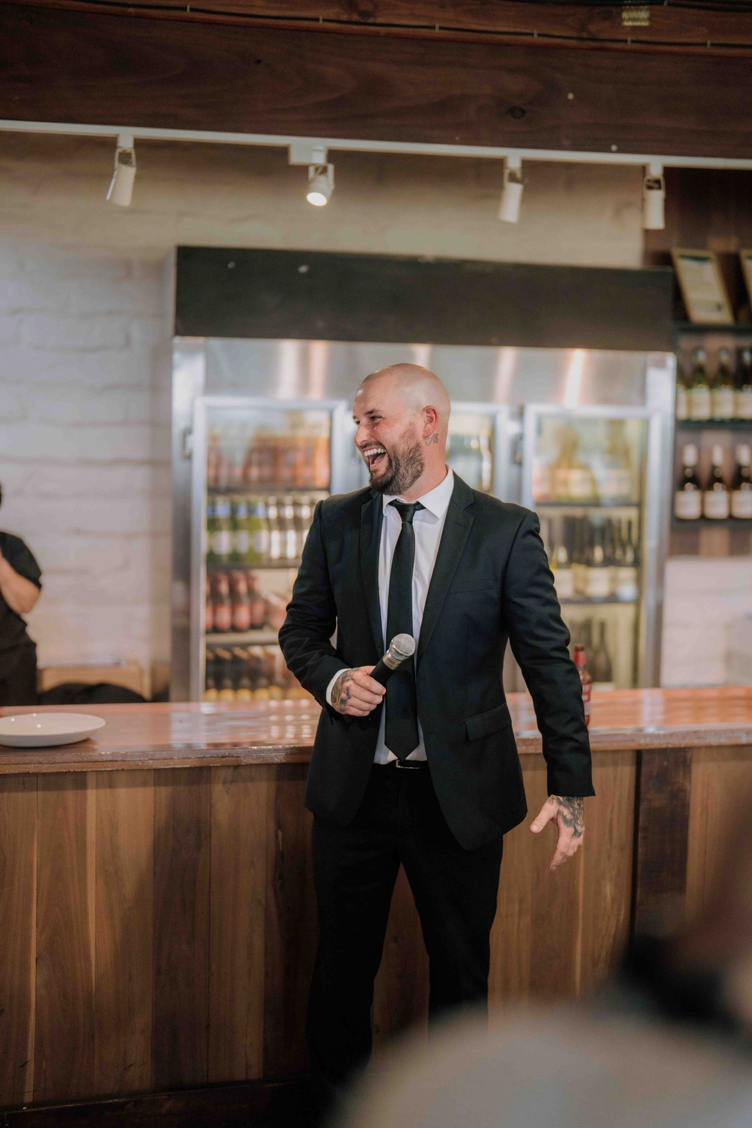 Bald man in dark suit smiles while holding microphone, giving speech at wedding reception near bar.