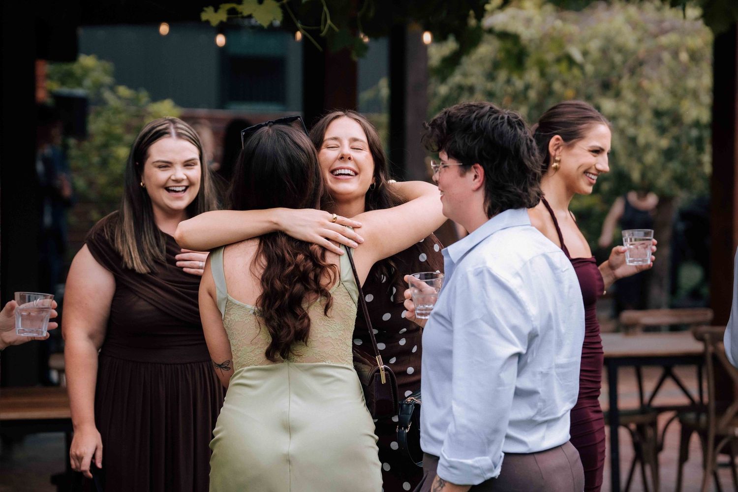 Group of smiling women in colorful dresses sharing a joyful hug at an outdoor wedding reception.