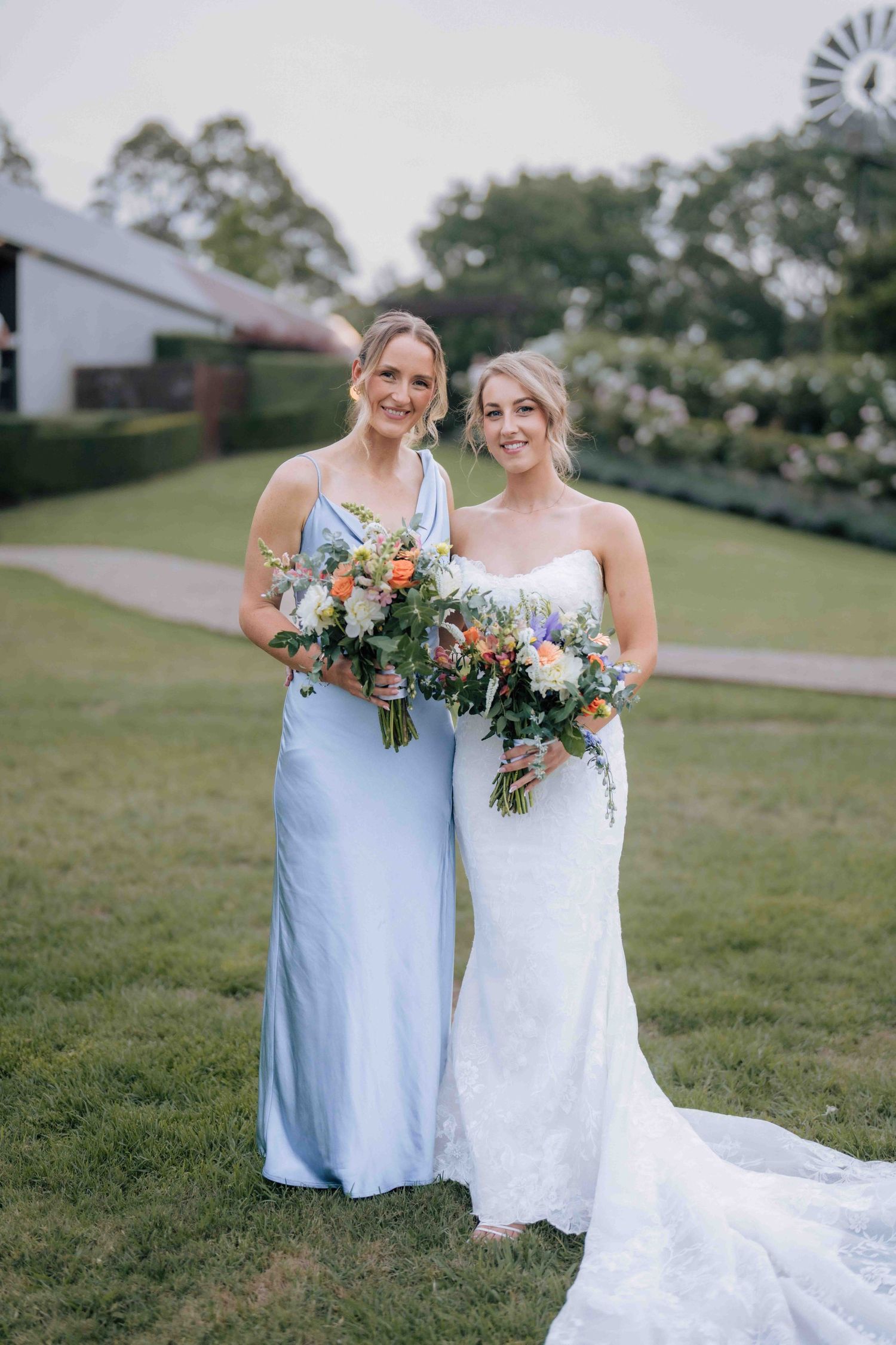 Bride in white lace gown poses with bridesmaid in light blue satin dress holding colorful floral bouquets outdoors.