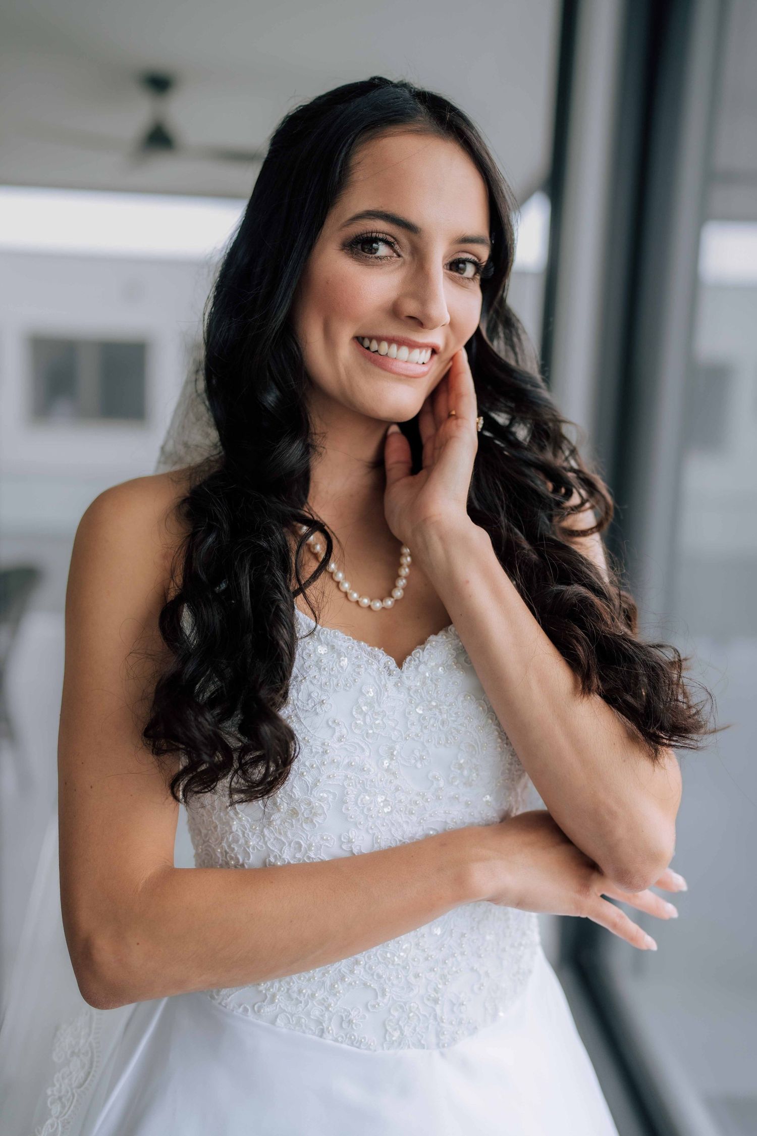 Beautiful dark-haired bride in white lace gown and pearls smiling softly by a bright window.