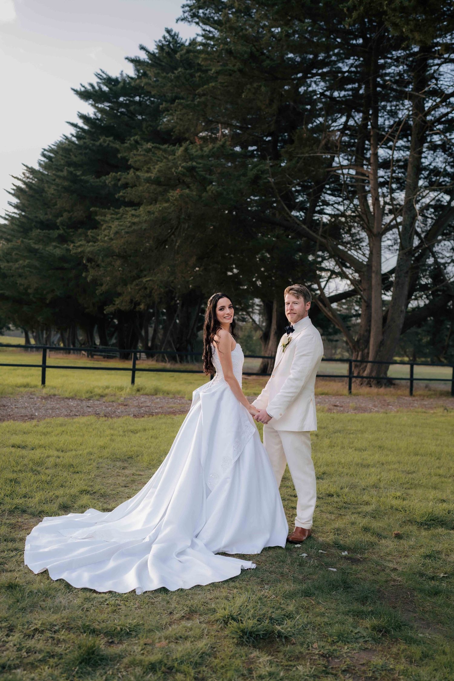 Elegant bride and groom hold hands outdoors, bride's long white gown trailing on green grass with tall trees behind.