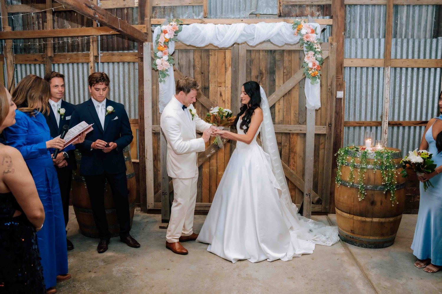 Bride and groom exchange vows under floral arch in rustic barn wedding ceremony with guests watching.