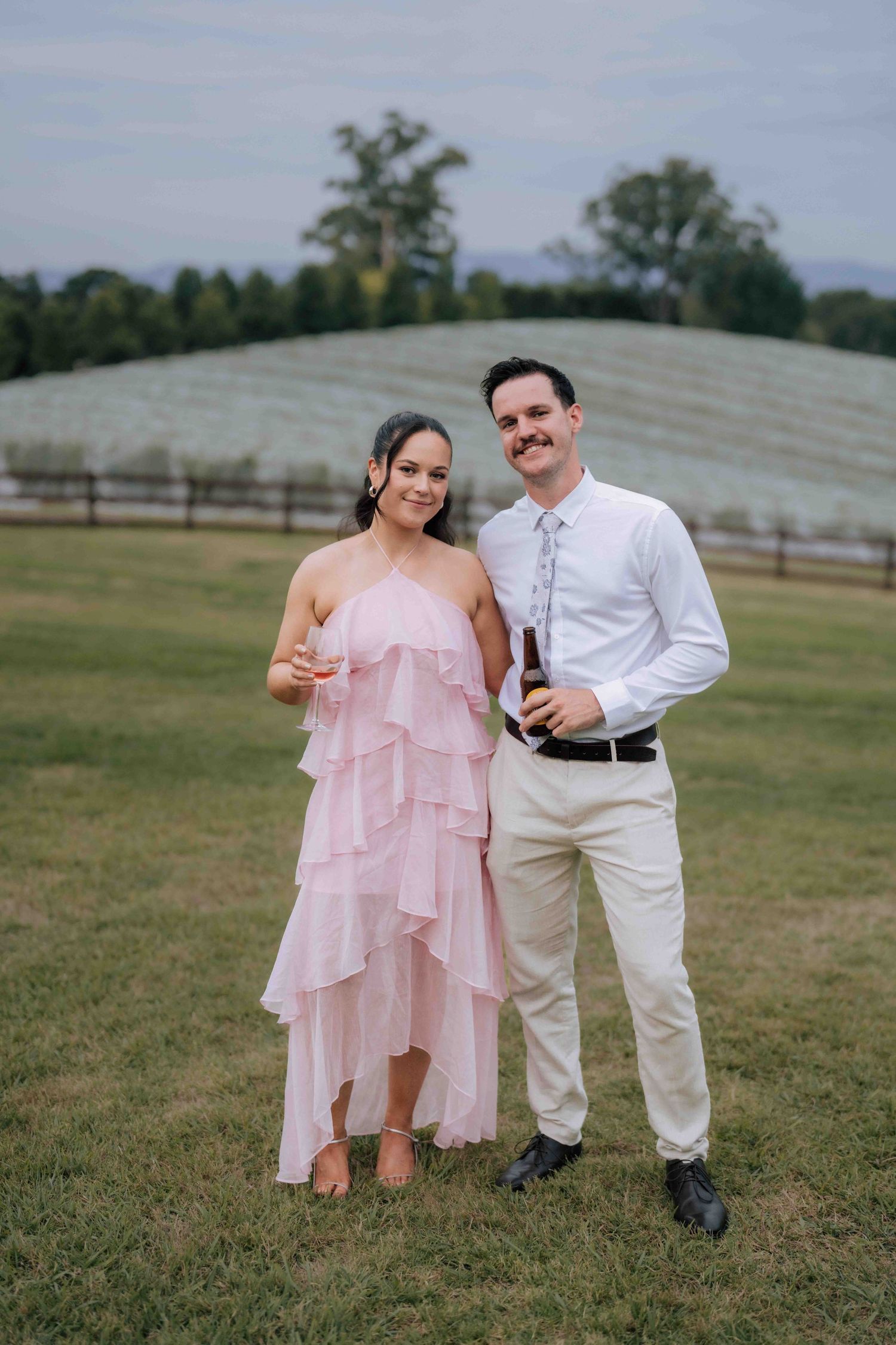 Smiling couple posing outdoors at a wedding, woman in pink ruffled dress, man holding a beer bottle.