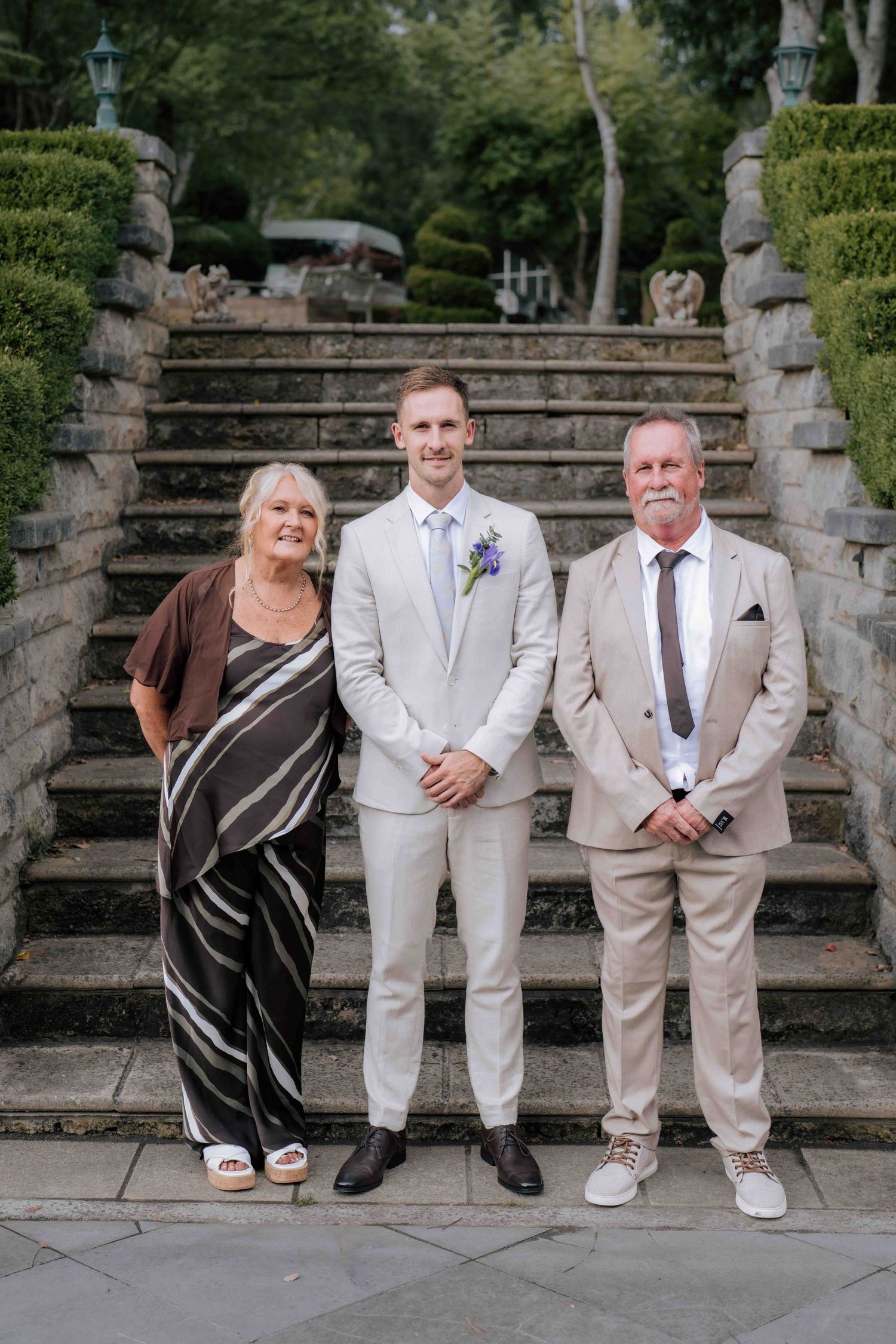 Groom in cream suit poses with parents on stone steps at outdoor wedding venue.
