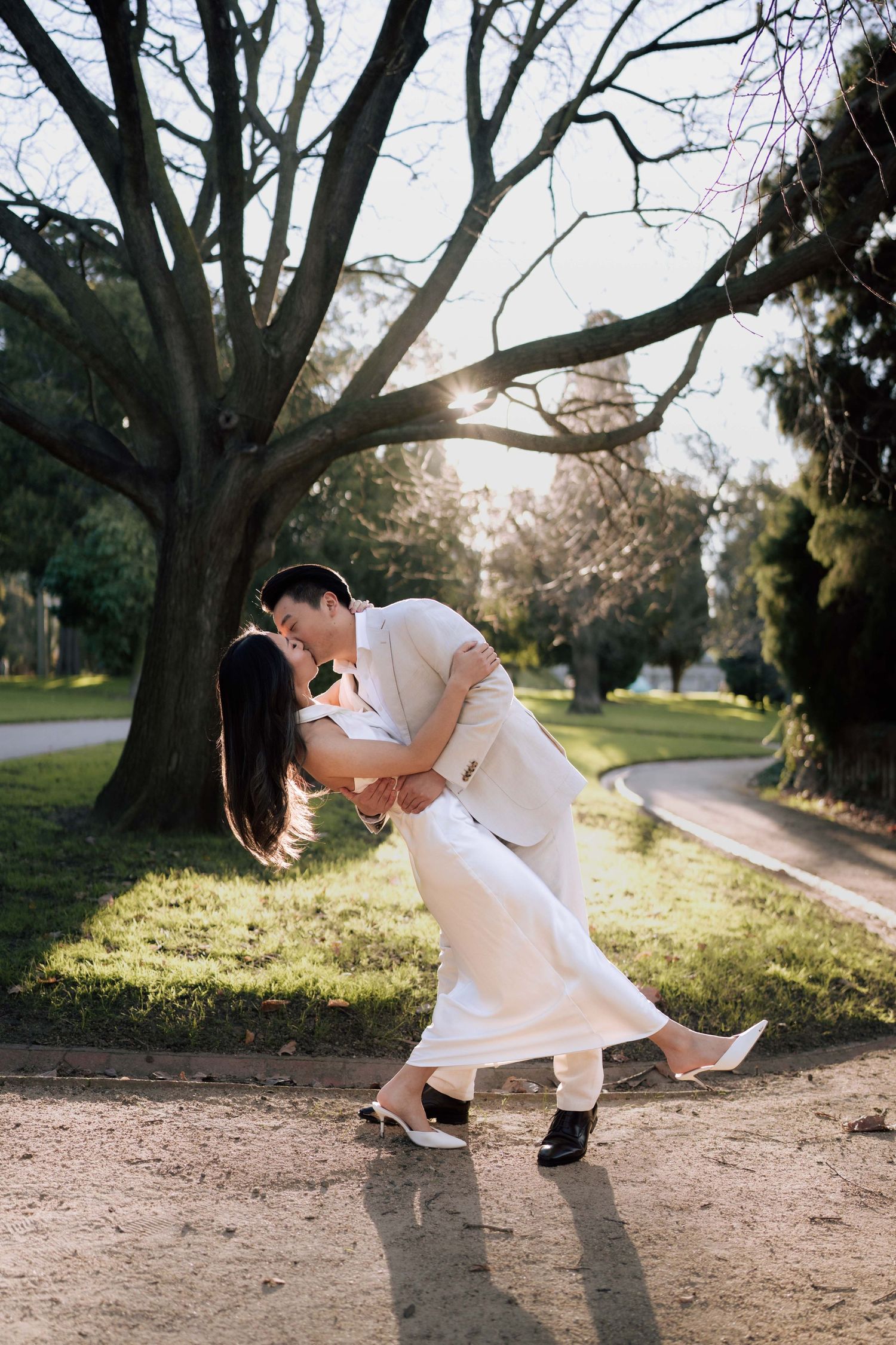 Asian couple in matching white outfits share a romantic dip kiss on a sunlit park path with bare trees.