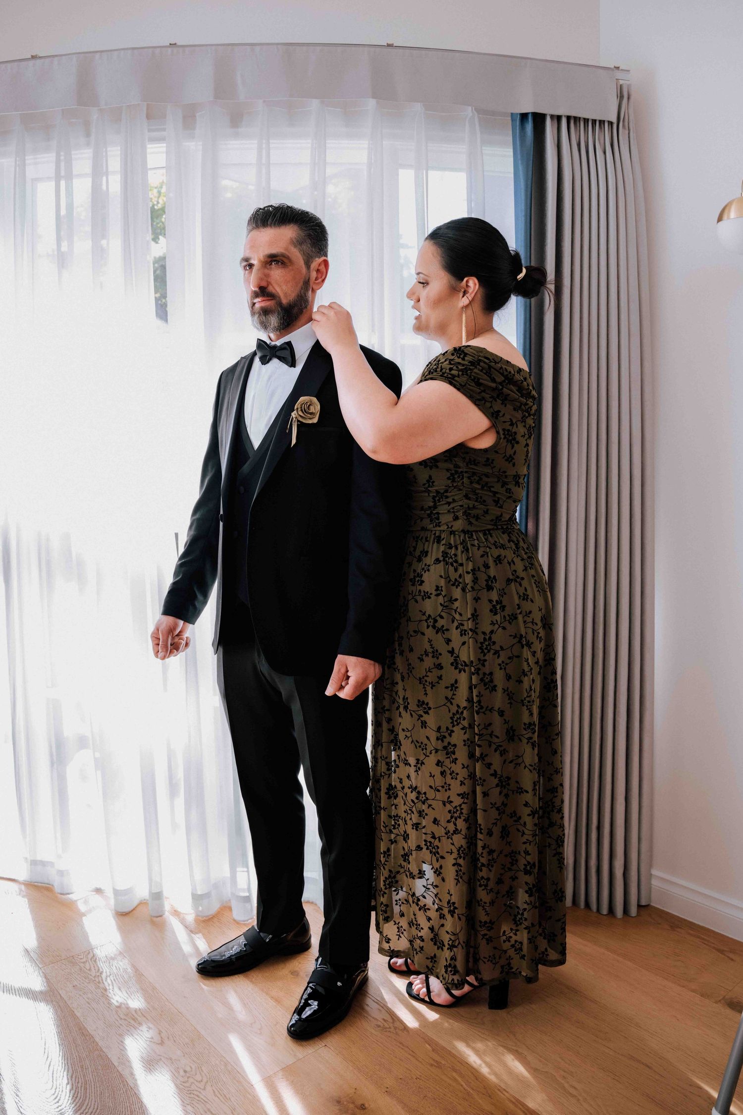 Groom in black tuxedo with bow tie gets boutonniere pinned by woman in gold floral gown at wedding.